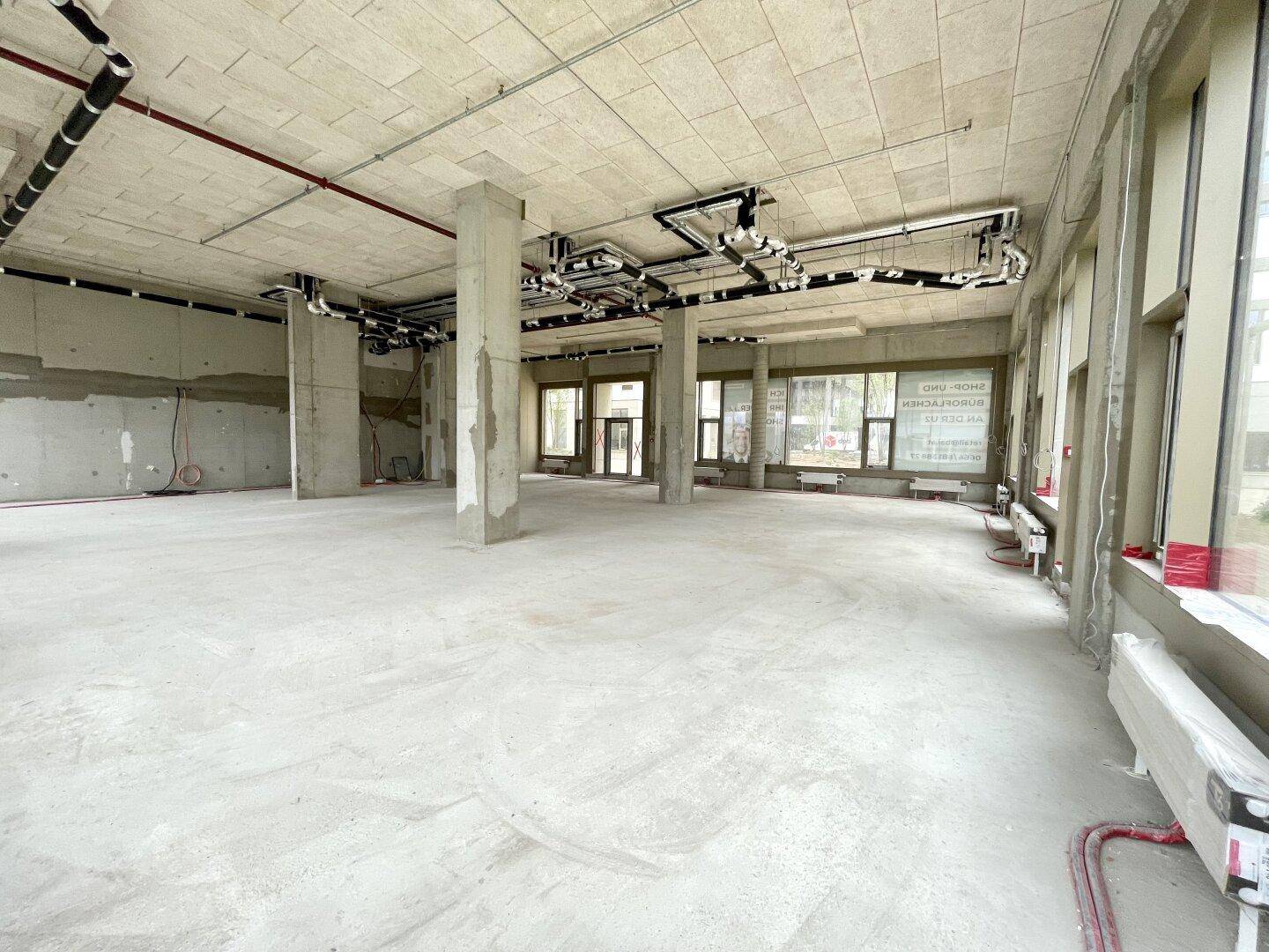 Weitläufiger Gewerberaum im Rohbauzustand mit Betonstützen und großen Fensterfronten.