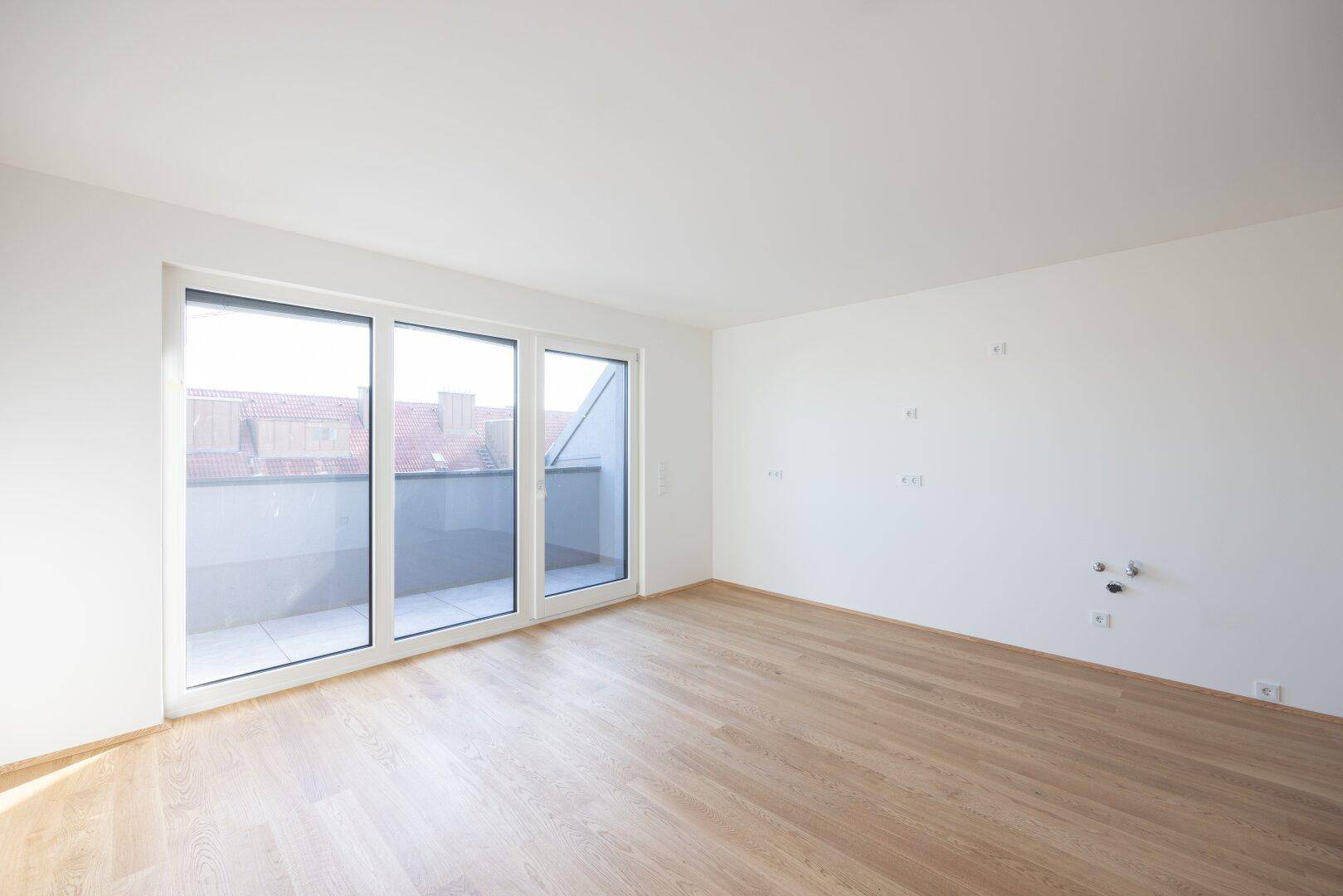 Heller, leerer Raum mit Holzboden und großen Fenstern, die Zugang zu einem Balkon bieten.