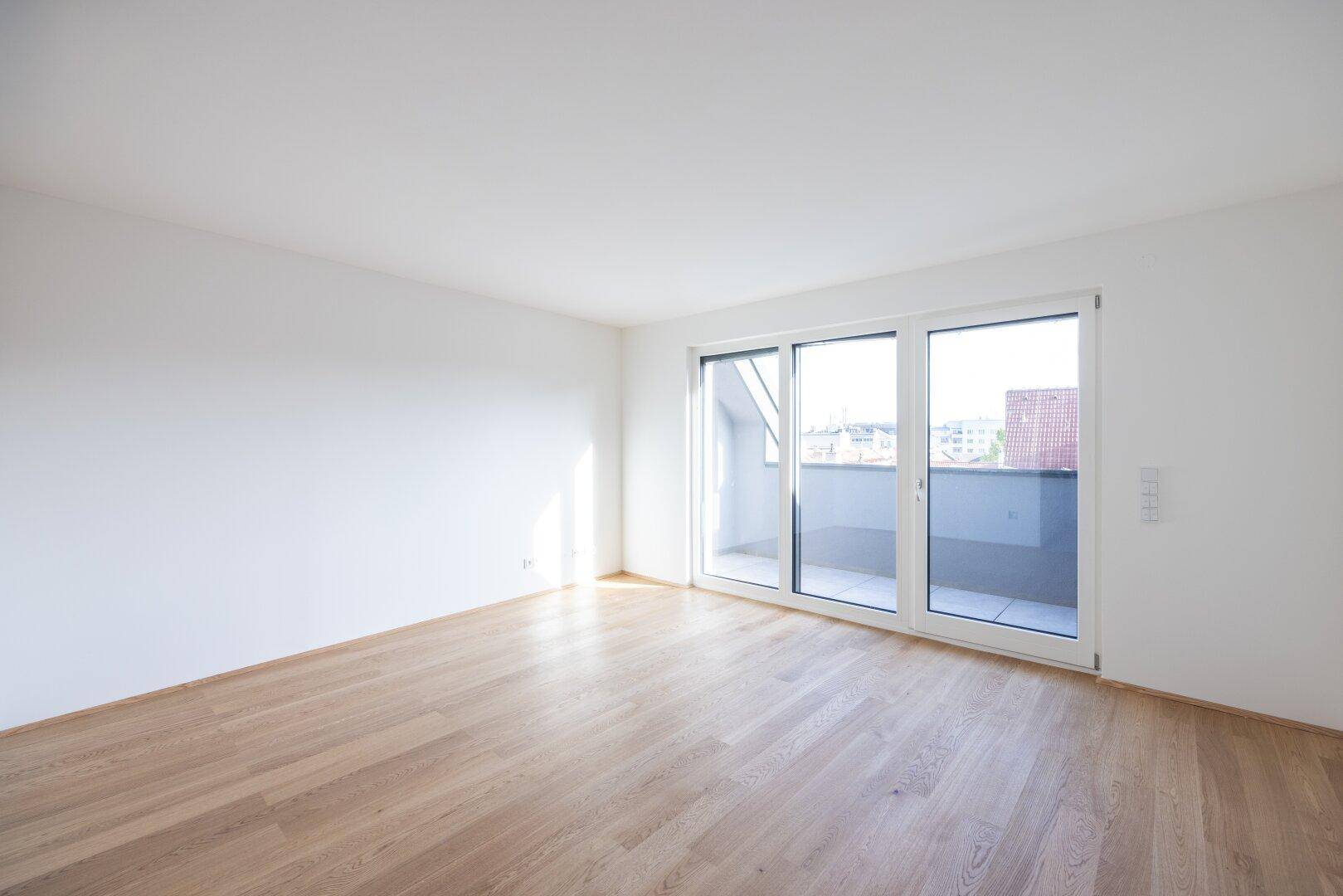 Geräumiger, unmöblierter Raum mit Holzboden und breiter Fensterfront, die viel Tageslicht hereinlässt.