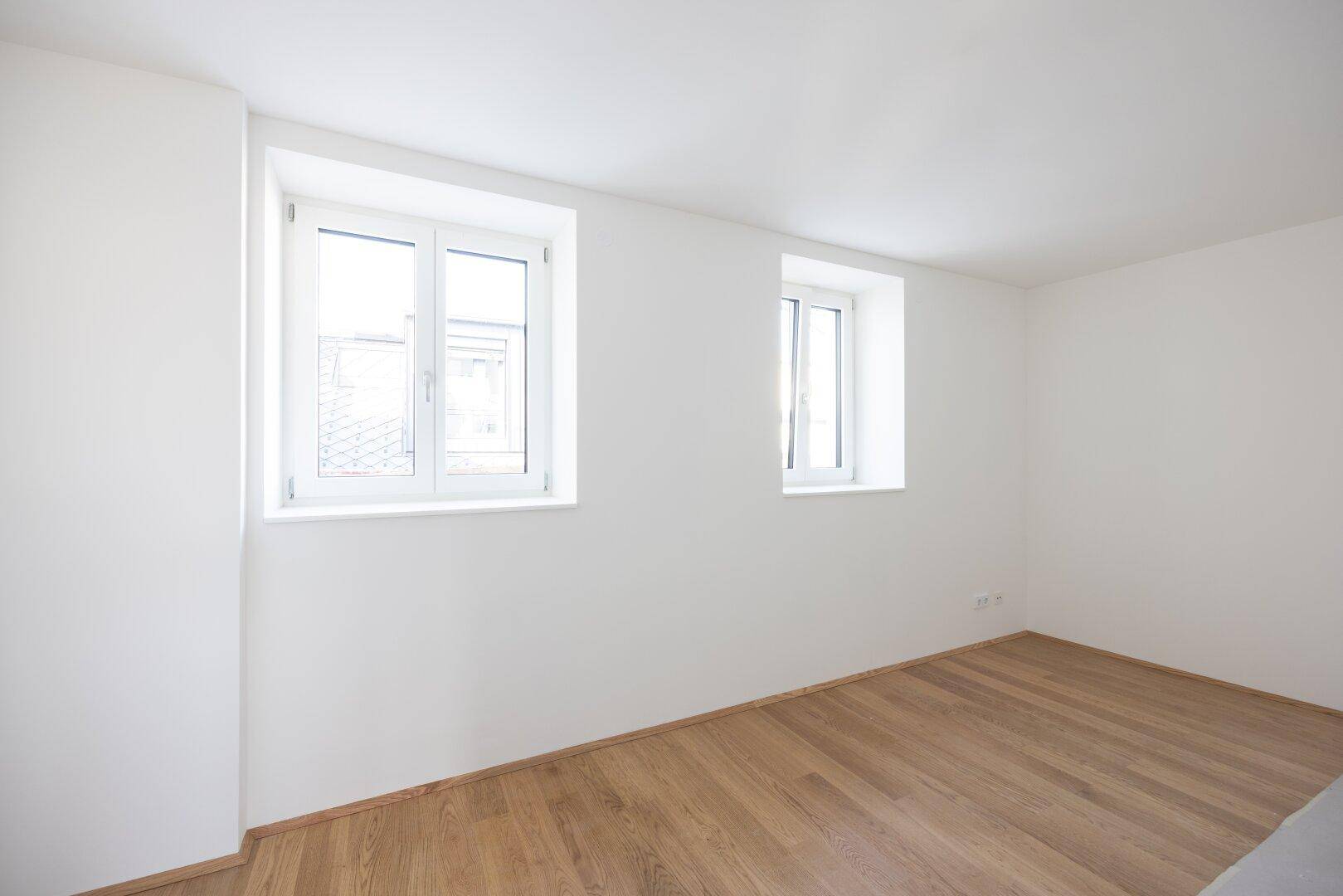 Unmöbliertes Schlafzimmer mit Holzboden und zwei Fenstern, die viel Tageslicht bieten.