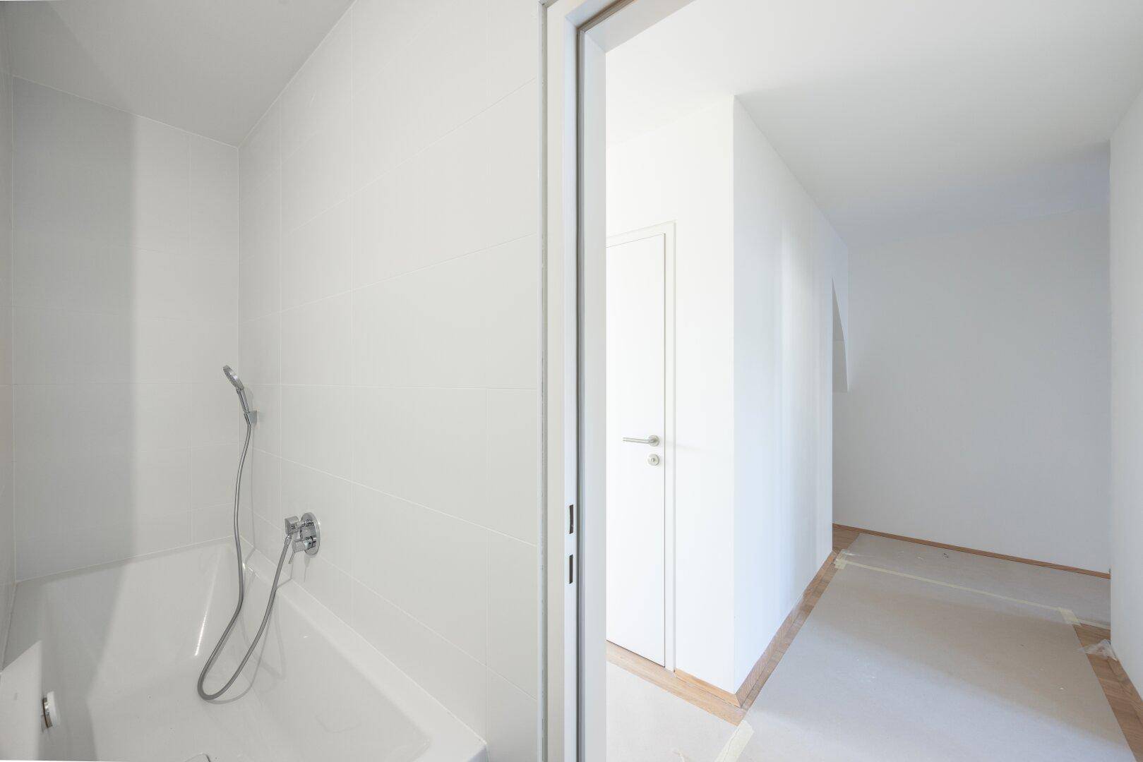 Modernes Badezimmer mit Badewanne und weißen Fliesen, angrenzend an einen hellen Flur.