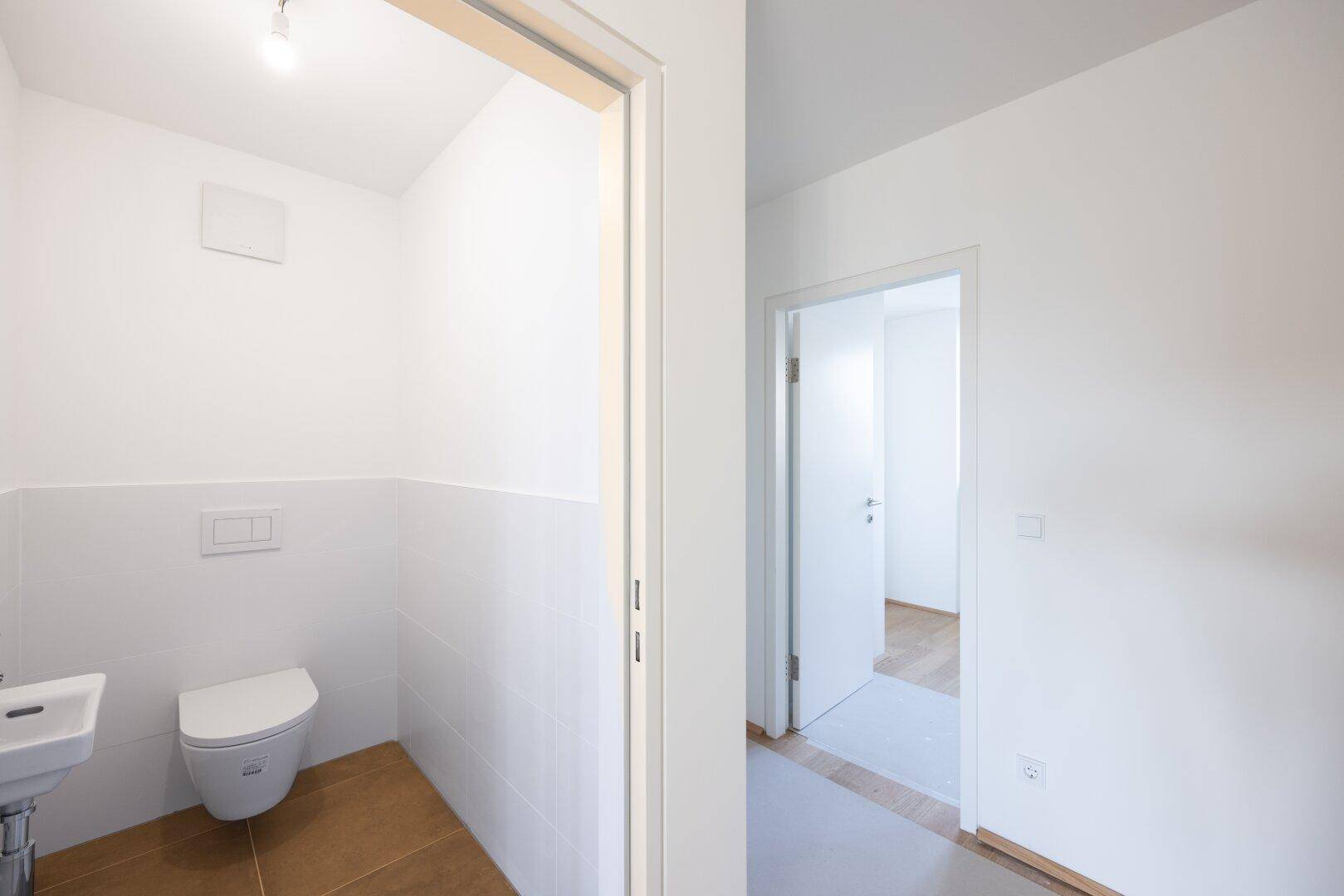 Helles Gäste-WC mit Wand-WC, kleinem Waschbecken und braunen Bodenfliesen.