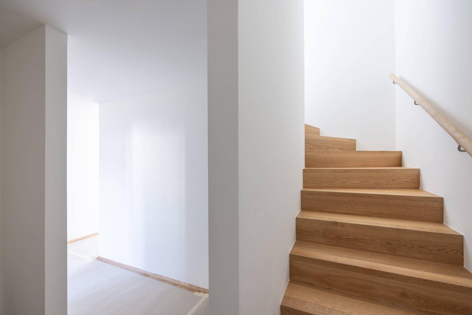 Holztreppe mit Geländer, die zu den oberen Etagen führt, in einem hellen Treppenhaus.