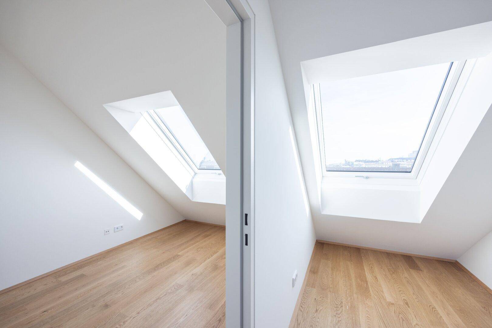 Zwei helle Räume im Dachgeschoss mit Holzboden und großen Dachfenstern, die viel Tageslicht hereinlassen.
