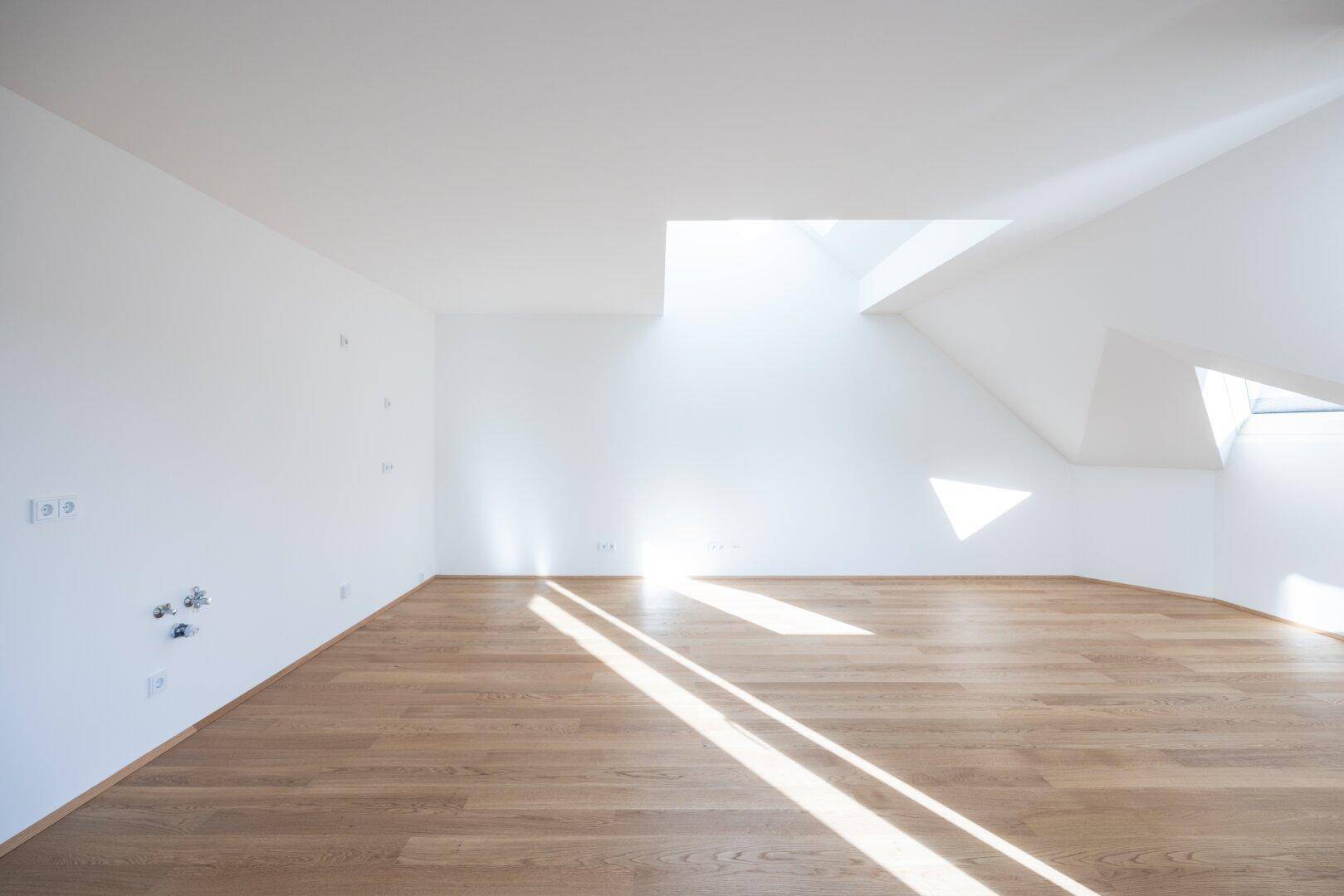 Heller, leerer Raum mit Holzboden und schrägen Wänden, durchflutet von Sonnenlicht durch Dachfenster.