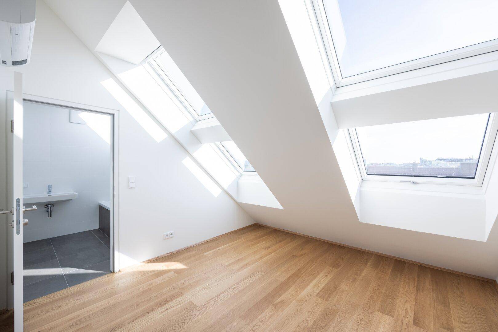 Heller Raum im Dachgeschoss mit mehreren Dachfenstern, Holzboden und Zugang zu einem Badezimmer.