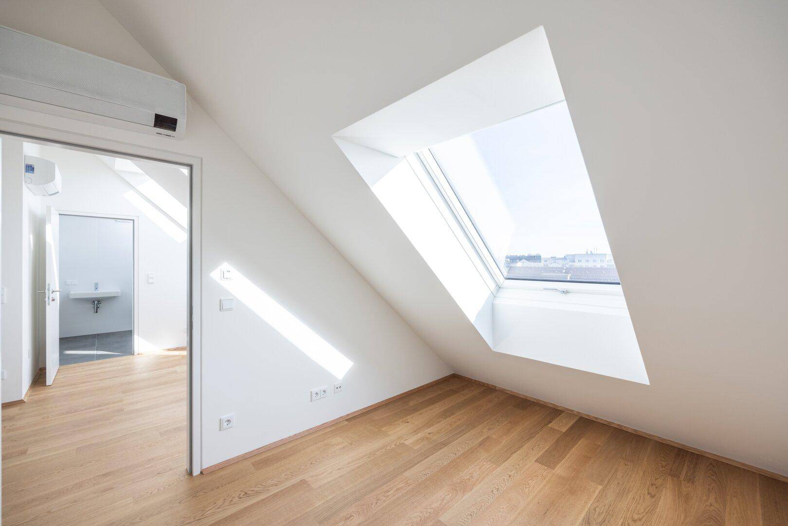 Heller Raum im Dachgeschoss mit Holzboden, Klimaanlage und großem Dachfenster, führt zu einem Bad.