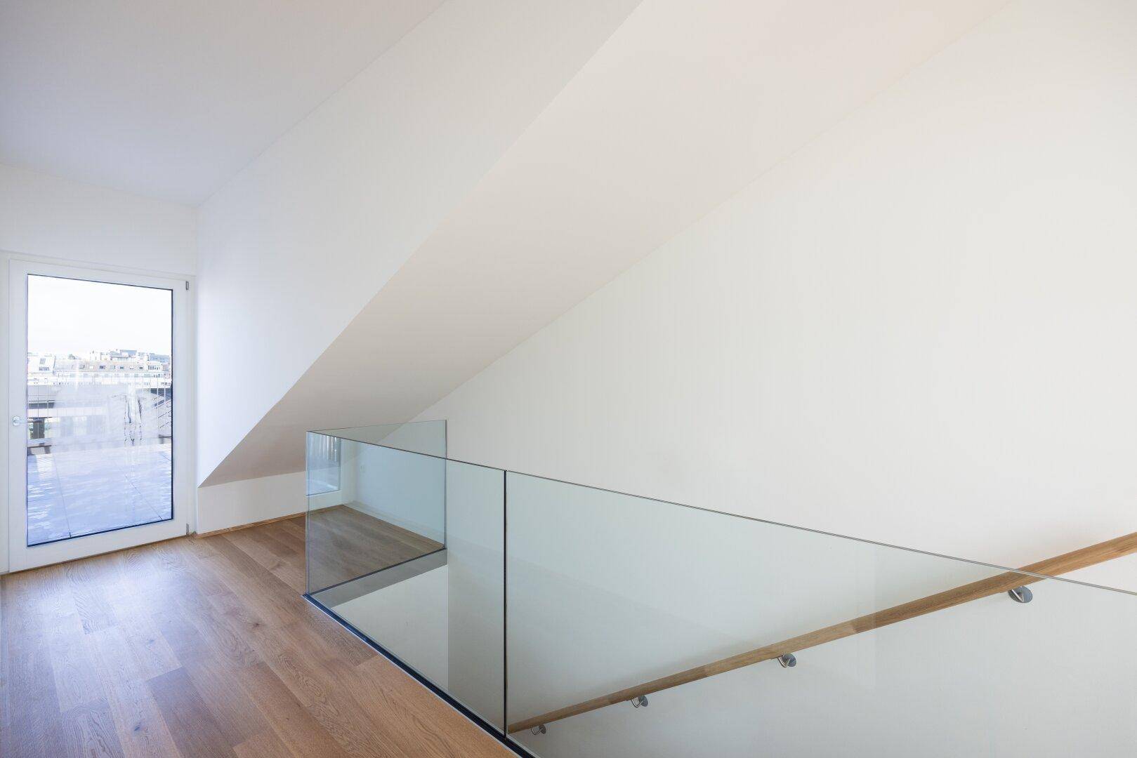 Moderner Treppenaufgang mit Glasgeländer und Holzboden, führt zu einem hellen Raum mit Terrassenzugang.