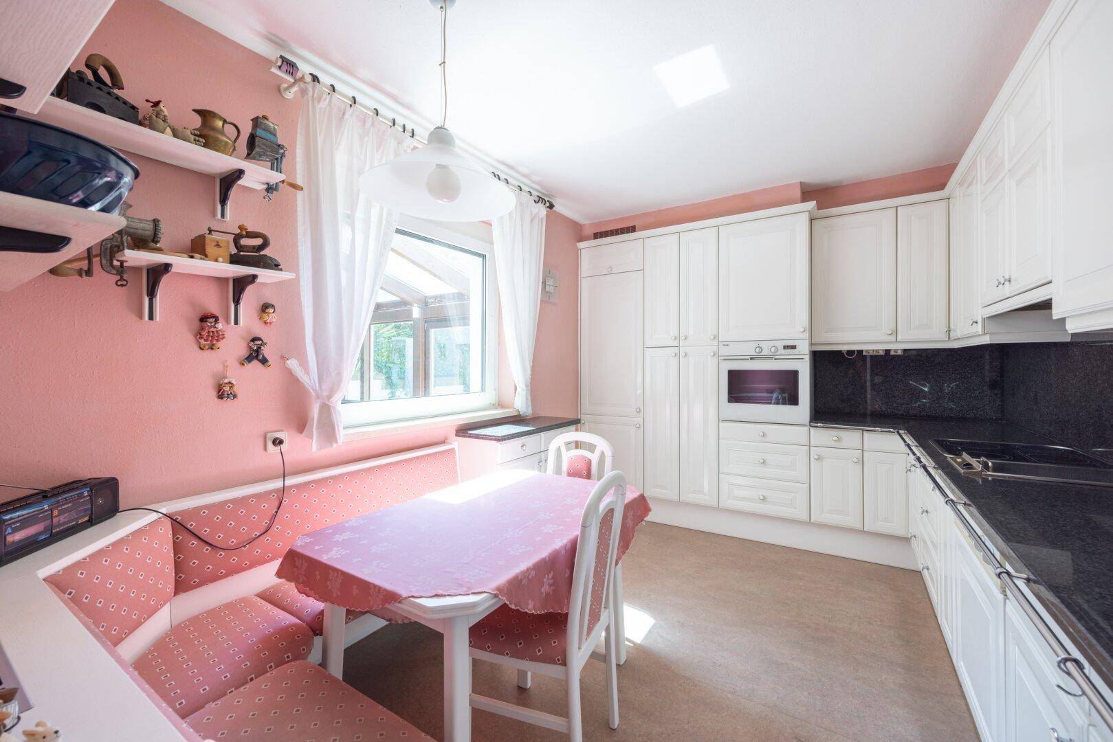 Gemütliche Wohnküche mit weißen Schränken, rosa Wänden und einem Essbereich am Fenster.
