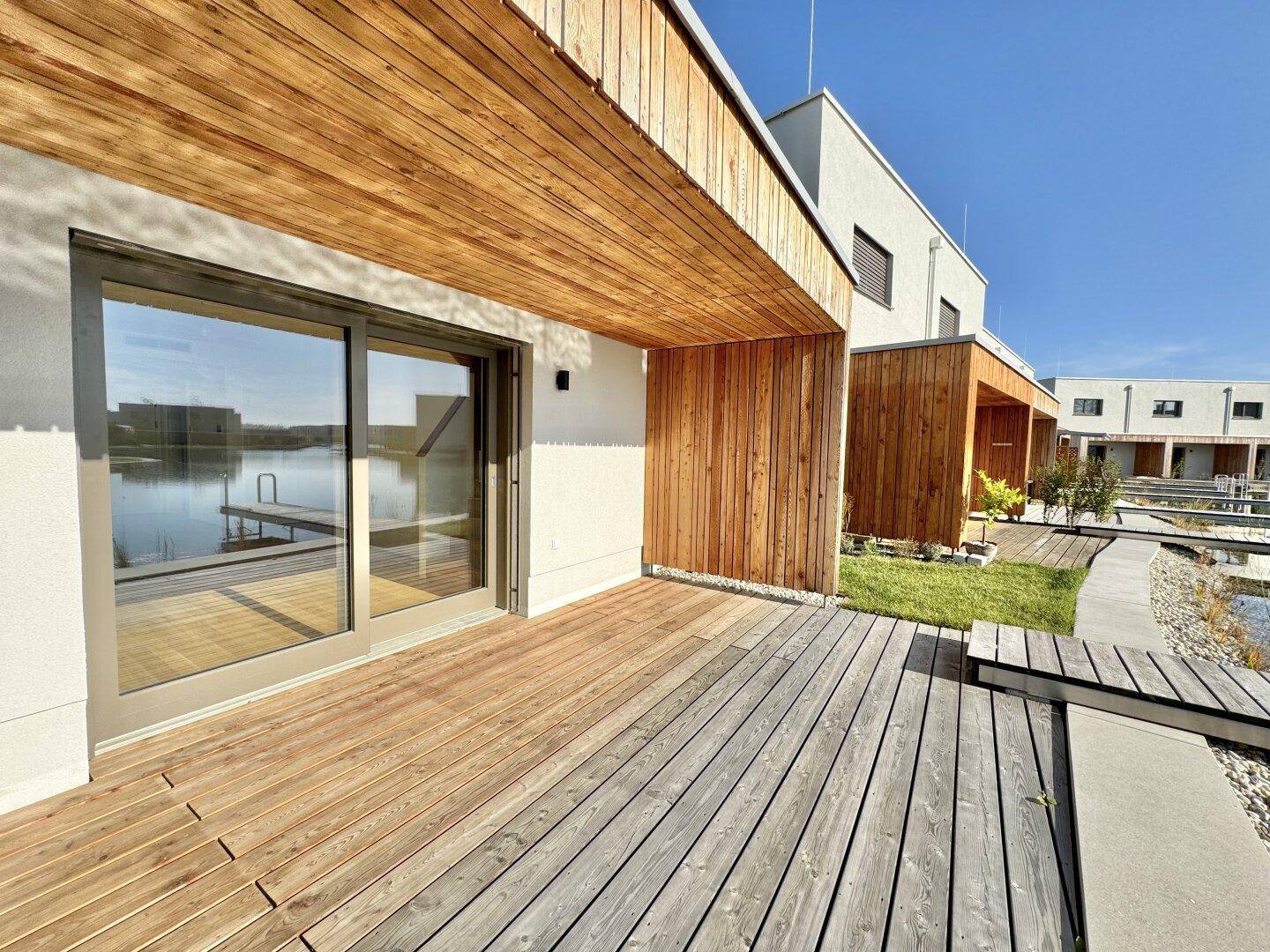 Große Holzterrasse mit Zugang zum Innenbereich und Blick auf den See, umgeben von moderner Architektur.