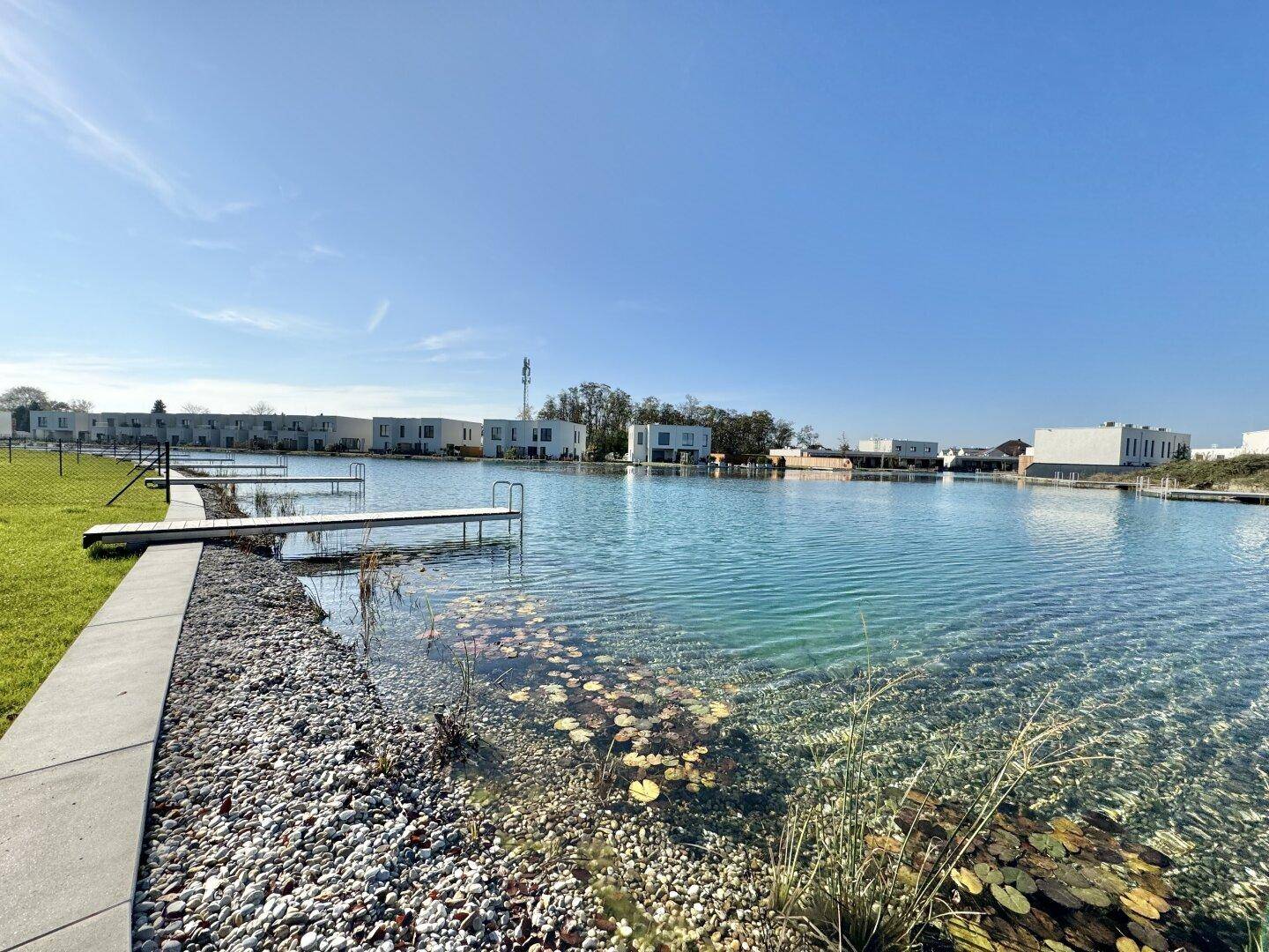 Klarer See mit Stegen und modernen Häusern am Ufer unter blauem Himmel.