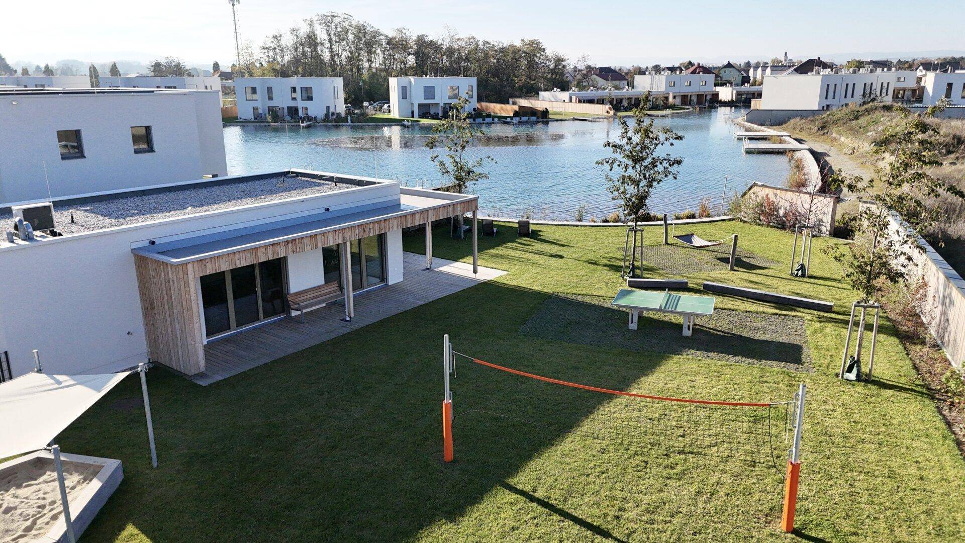 Gemeinschaftlicher Gartenbereich mit Rasen, Sandkasten und Volleyballnetz am Seeufer.