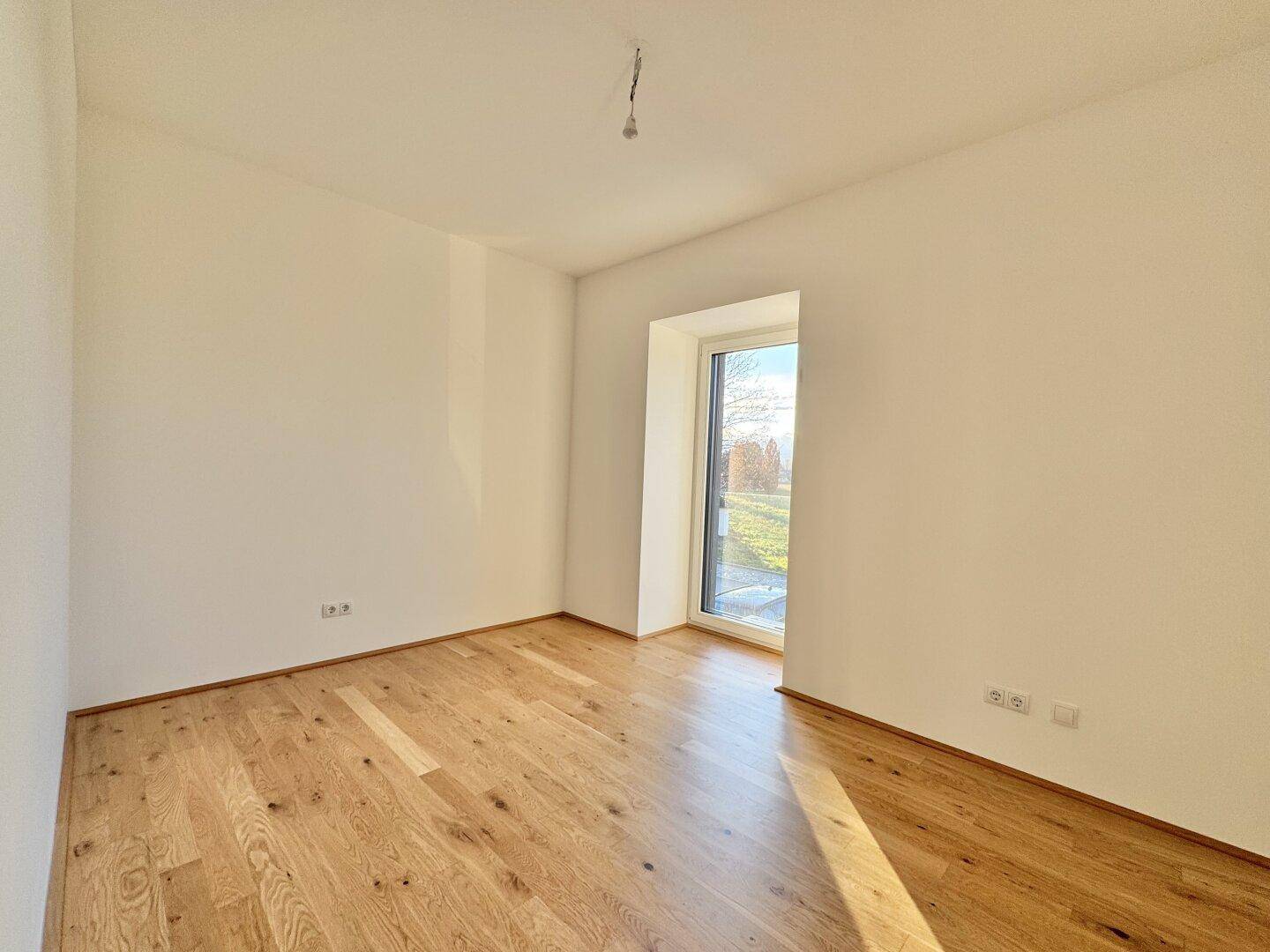 Helles Schlafzimmer mit Eichenparkettboden und einem hohen Fenster, das viel Sonnenlicht hereinlässt.