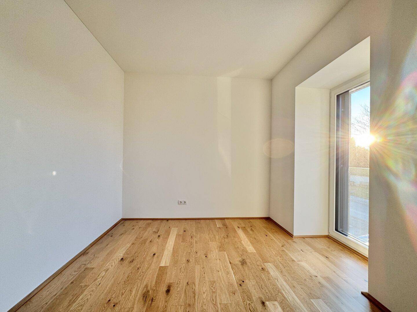 Lichtdurchflutetes Zimmer mit hellem Parkettboden und einem großen Fenster mit Sonneneinfall.