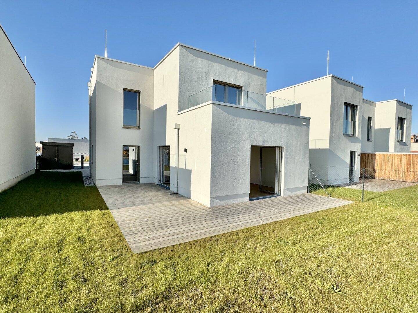 Moderne Neubau-Doppelhaushälfte mit Holzterrasse und Gartenbereich unter blauem Himmel.