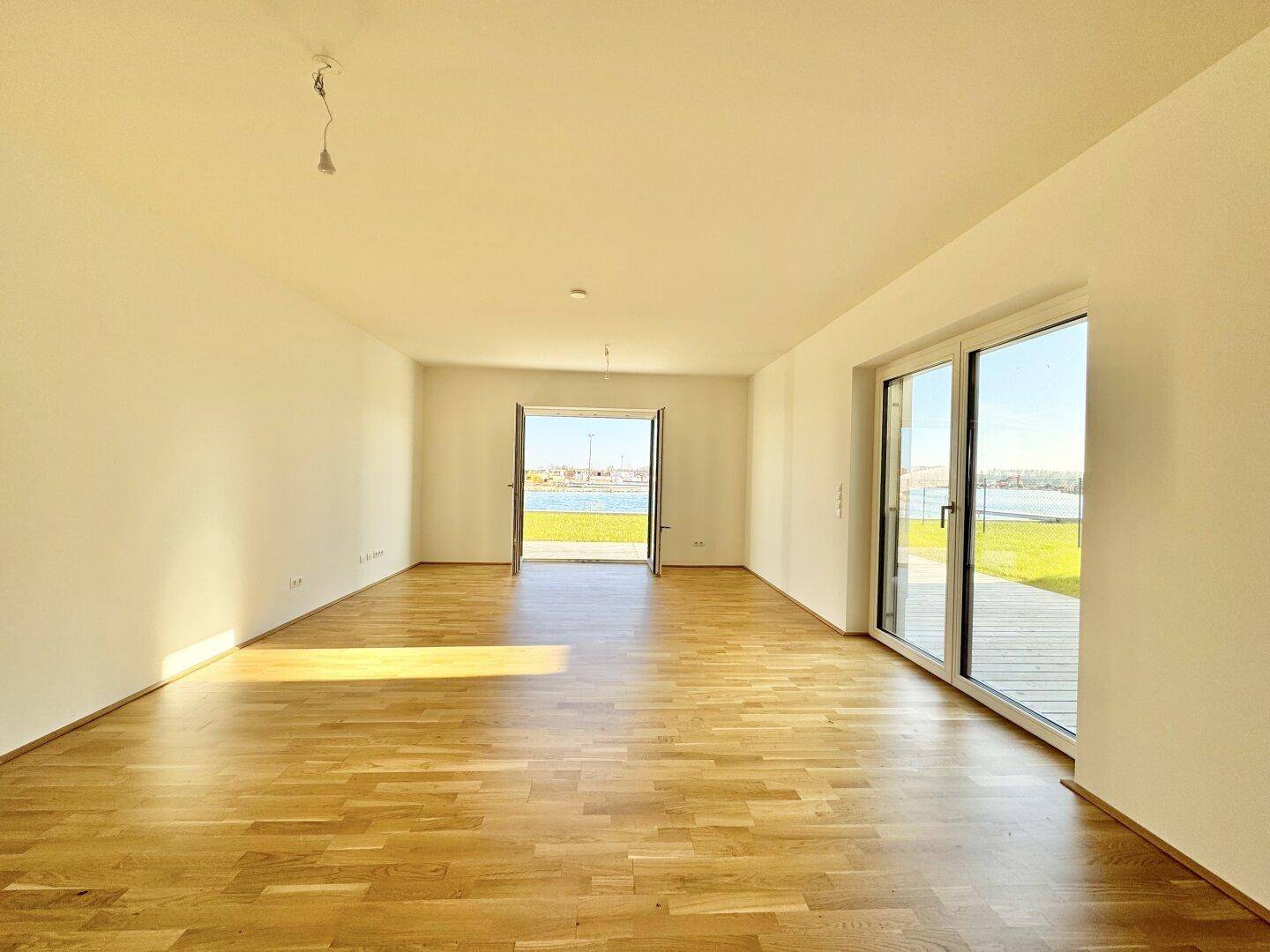 Helles, leeres Wohnzimmer mit Holzboden und direktem Zugang zur Terrasse mit Seeblick.