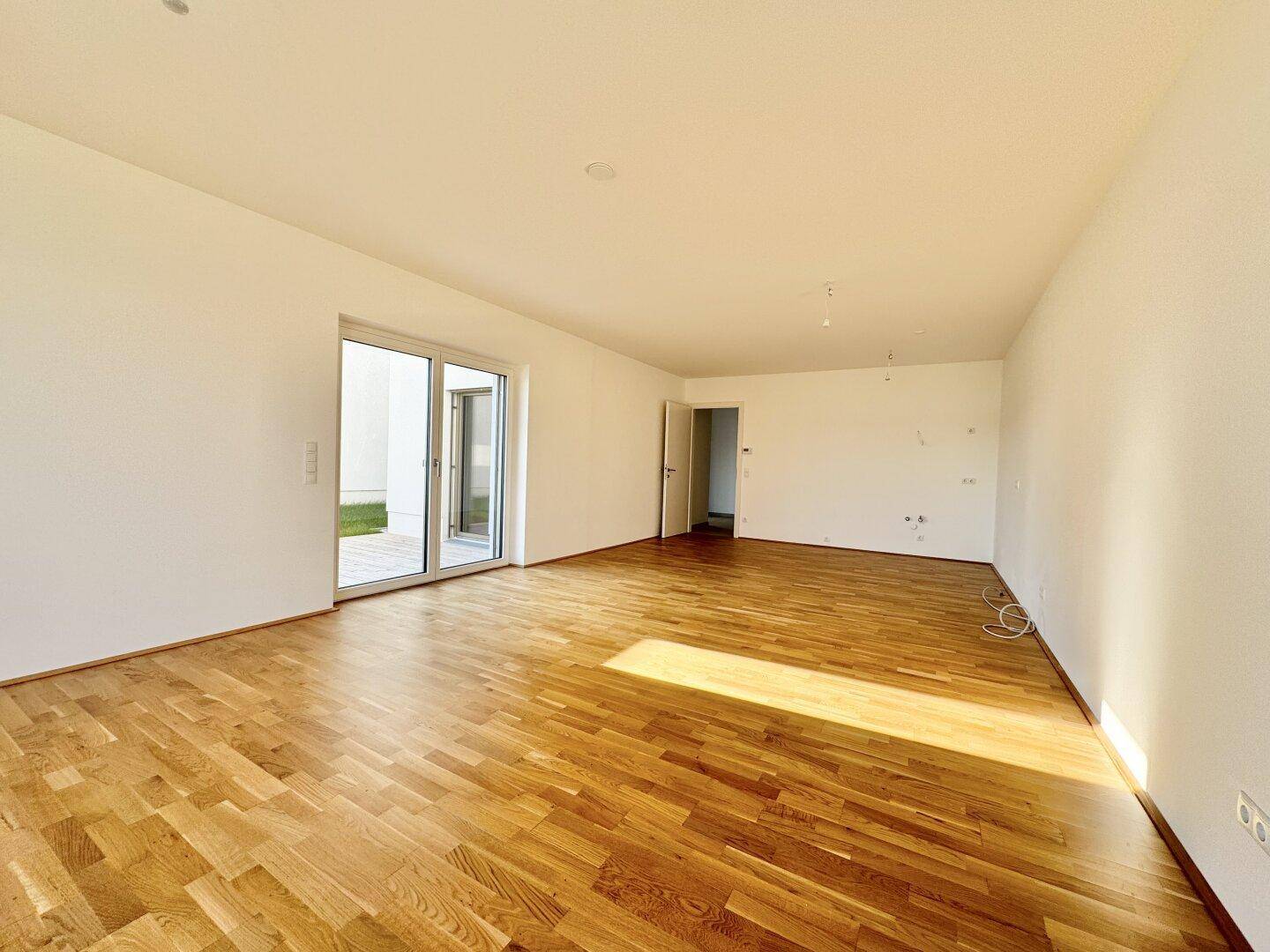 Geräumiges, leeres Wohnzimmer mit Parkettboden und Zugang zu einem kleinen Gartenbereich.