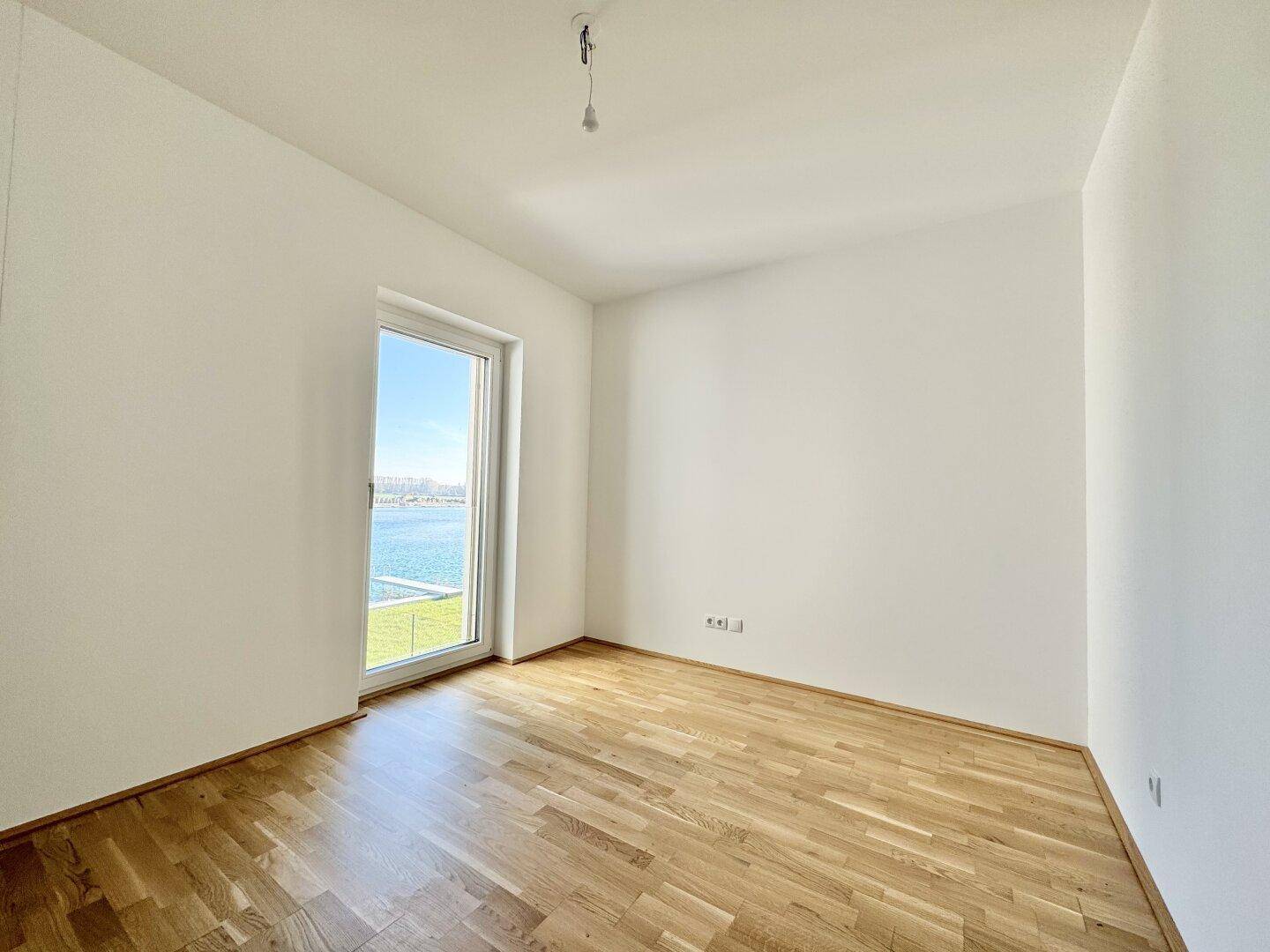 Leeres, helles Zimmer mit Holzboden und großem Fenster mit direktem Seeblick.