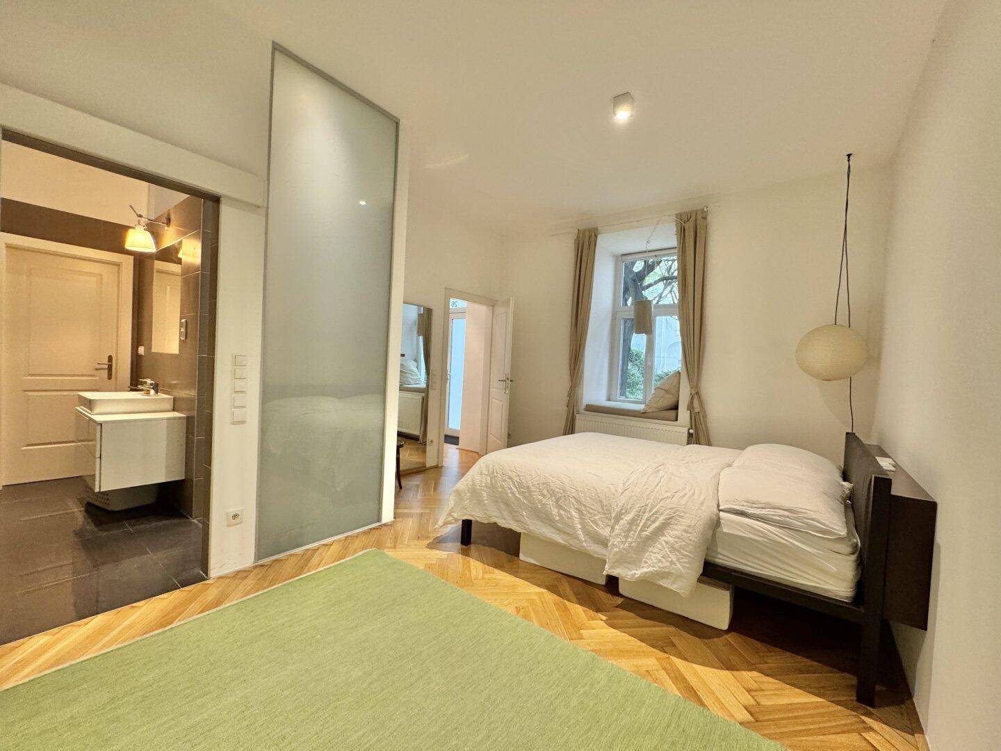 Schlafzimmer mit Parkettboden, großem Fenster und direktem Zugang zum Badezimmer.