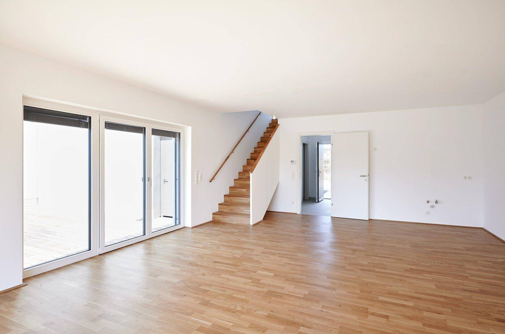 Geräumiger, leerer Wohnbereich mit großen Fenstern, Holzboden und offener Holztreppe.