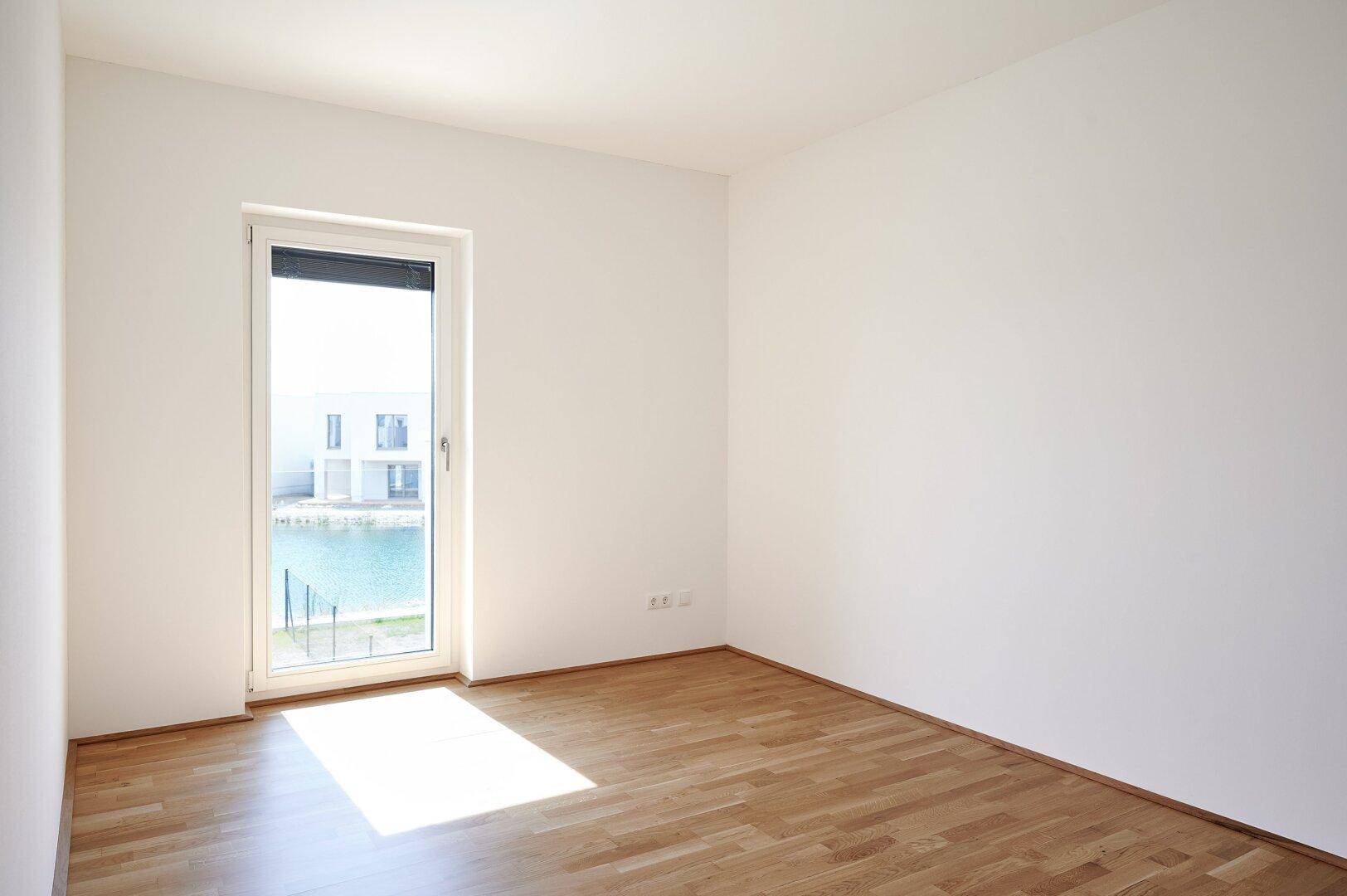 Leeres, helles Schlafzimmer mit Holzboden und einem Fenster mit Blick auf die Wasserfläche.