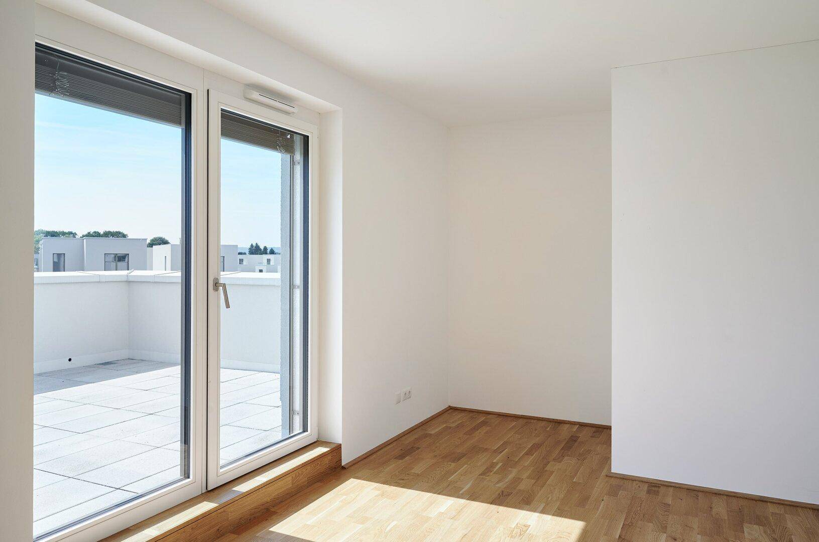 Leeres Schlafzimmer mit Holzboden und großen Fenstern, die Zugang zu einer Terrasse bieten.