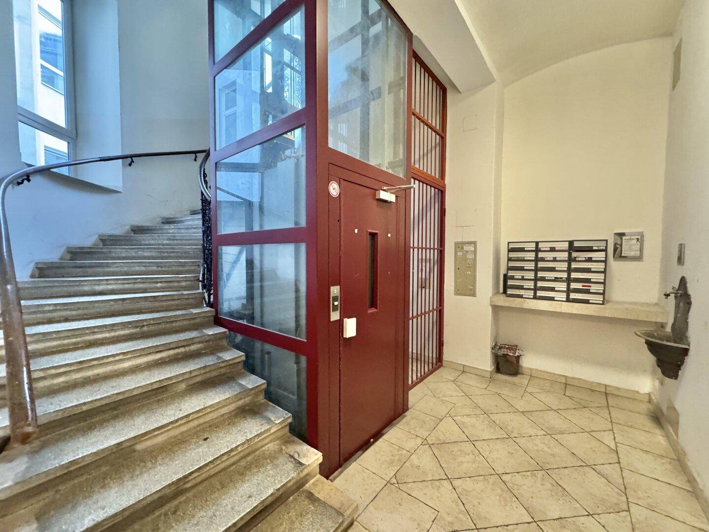 Historisches Treppenhaus mit modernem Glasaufzug und Marmorstufen, das die Stockwerke verbindet.