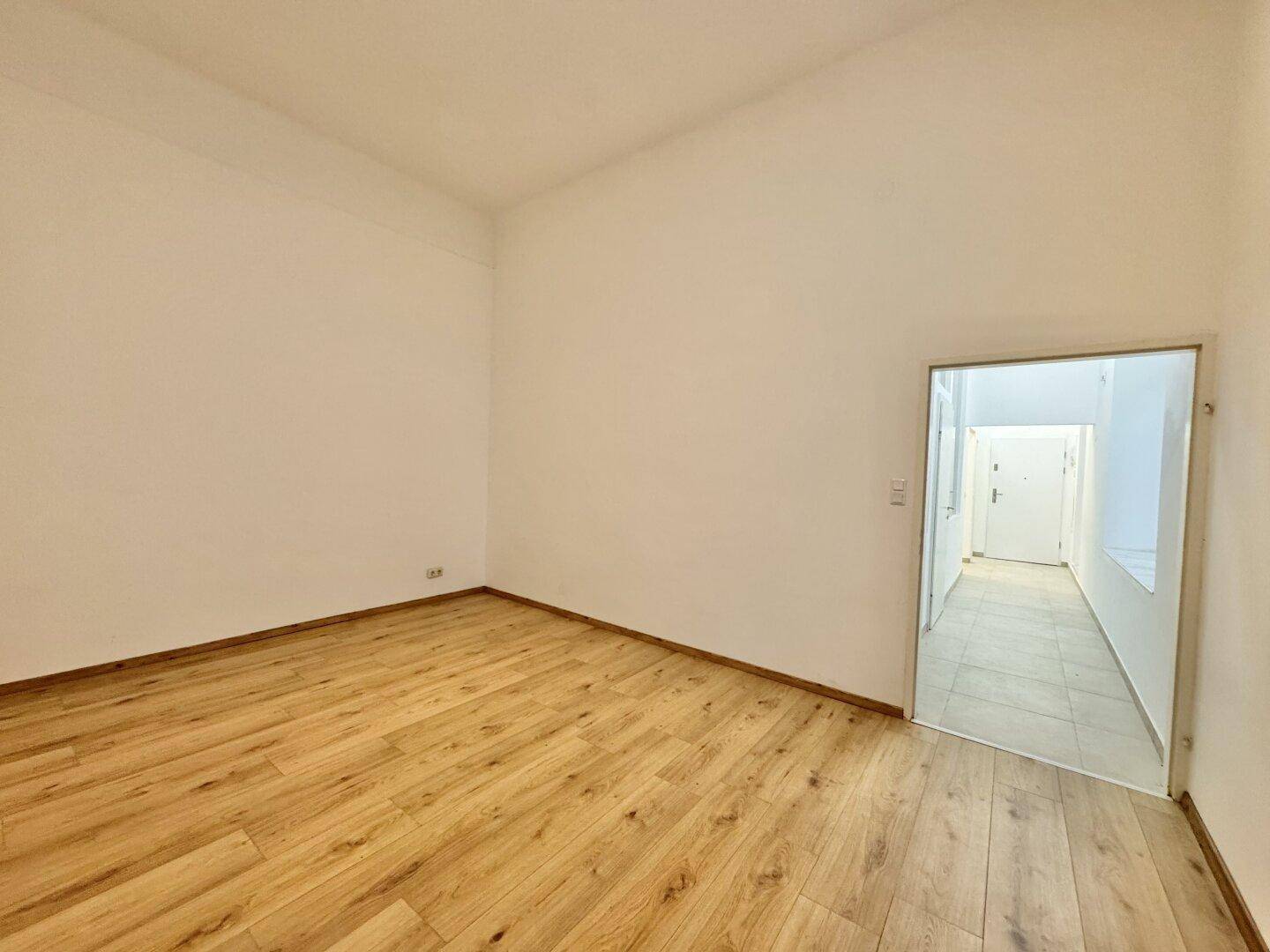 Heller, leerer Raum mit Holzboden und Zugang zu einem gefliesten Flur.