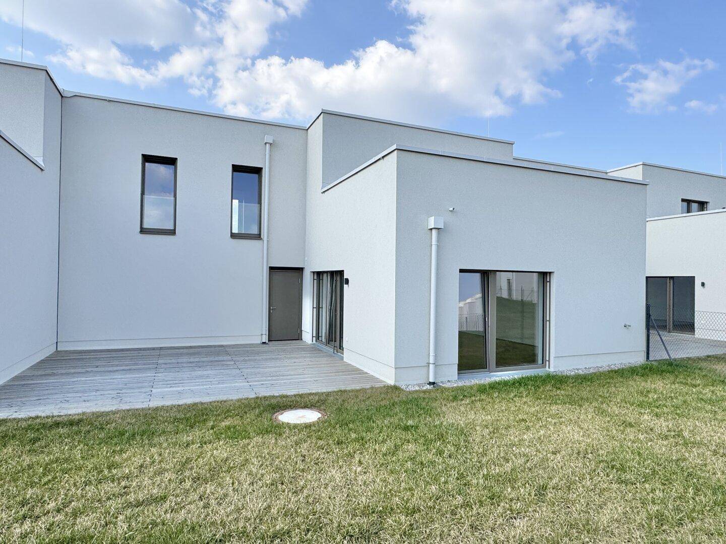 Rückseite eines modernen Hauses mit großer Holzterrasse und angrenzender Rasenfläche.