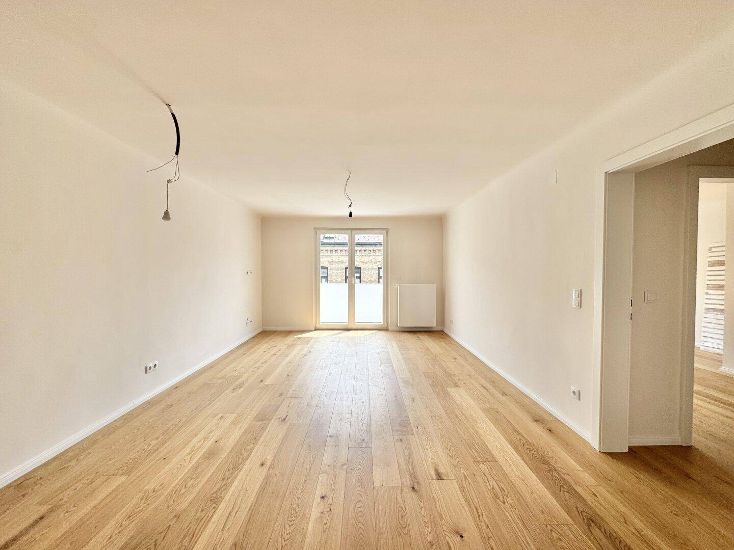 Heller, leerer Raum mit Holzboden und Zugang zu einem Balkon durch eine Fenstertür.