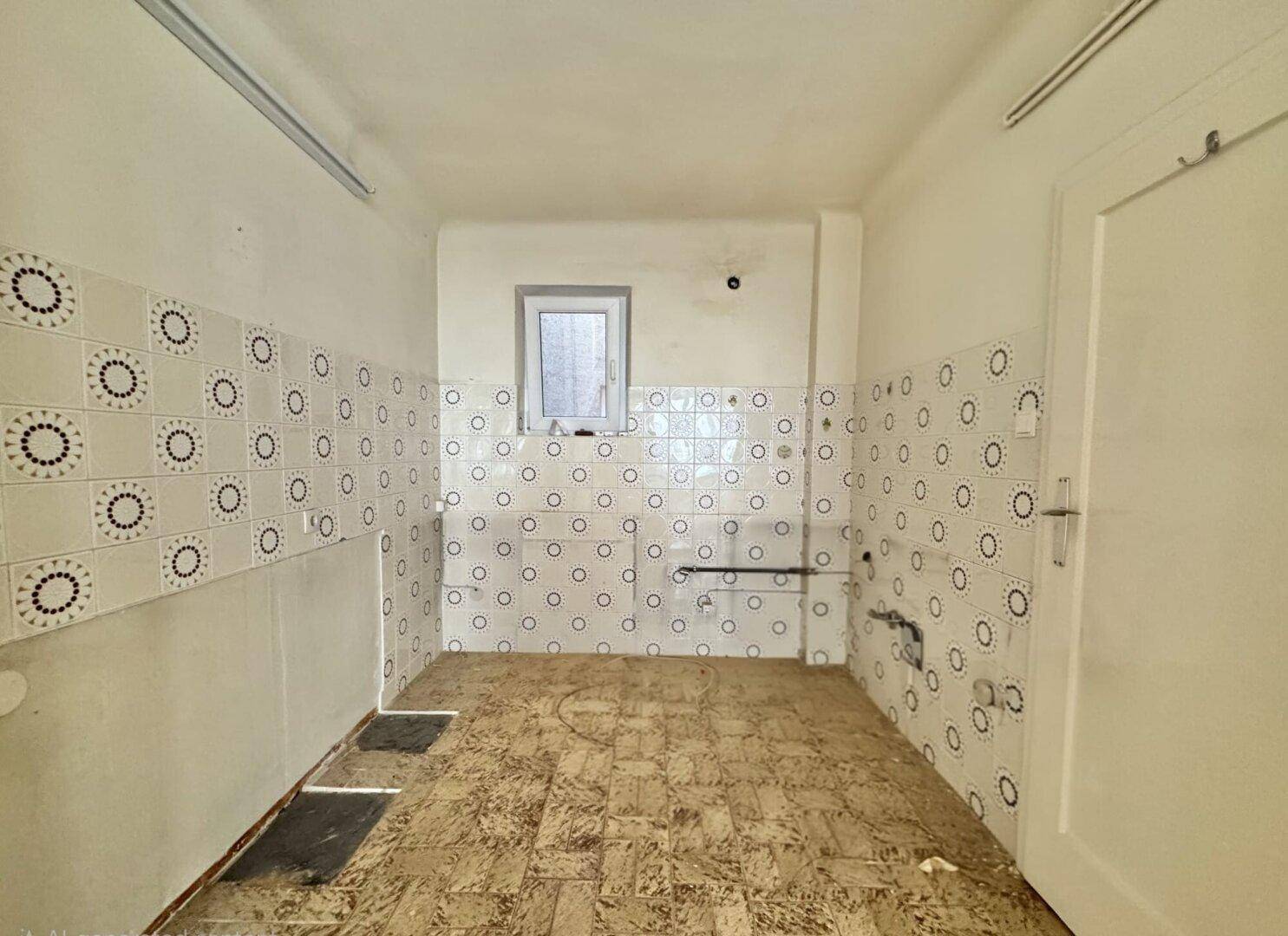 Leere Küche in sanierungsbedürftigem Zustand mit alten Fliesen, Fenster und sichtbaren Installationen.