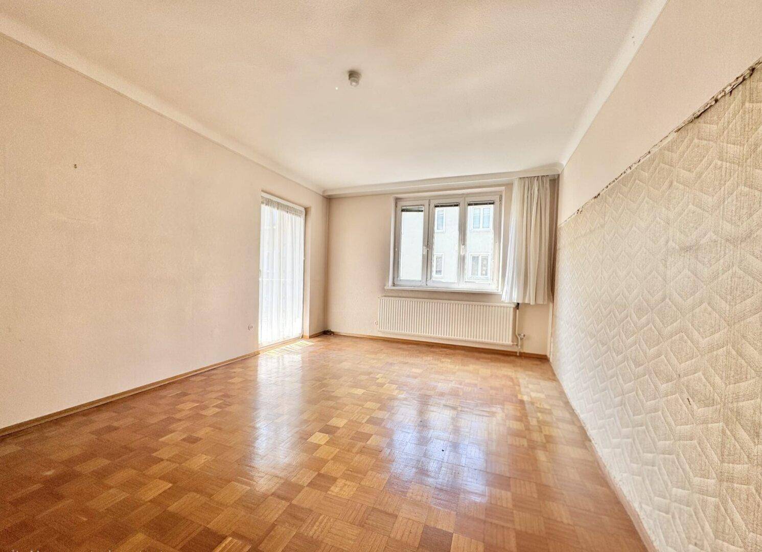 Leeres Zimmer mit Fischgrätparkett, Fenster und Zugang zu einem Außenbereich, in renovierungsbedürftigem Zustand.