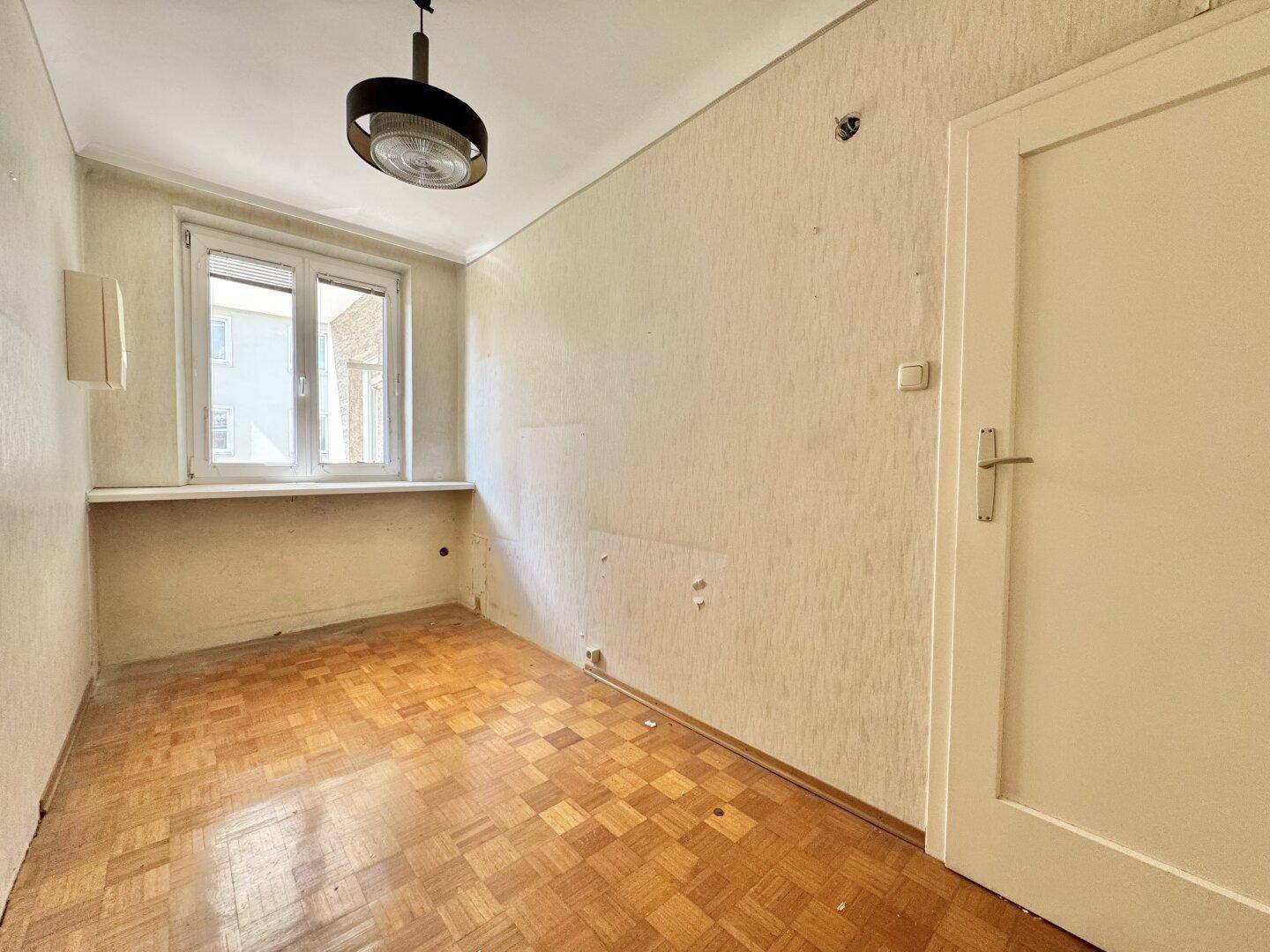 Kleines leeres Zimmer mit Parkettboden, Fenster und einfacher Beleuchtung, ideal als Kinderzimmer.