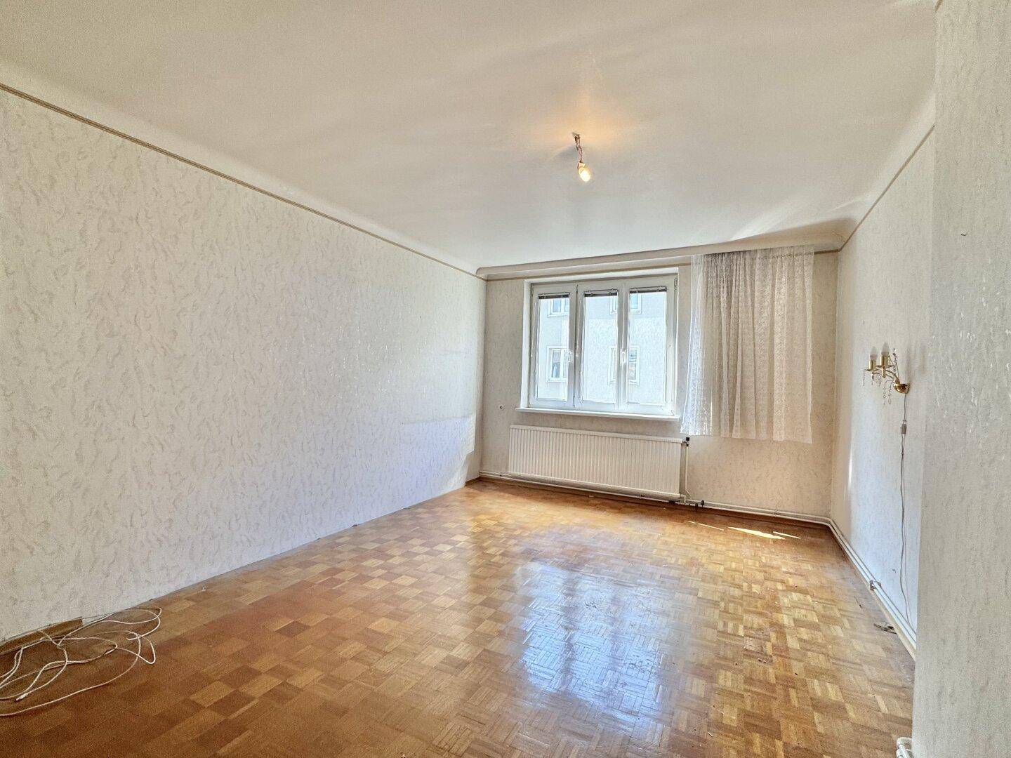 Leeres Zimmer mit Parkettboden, Fenster und Heizkörper, bereit für eine Renovierung.