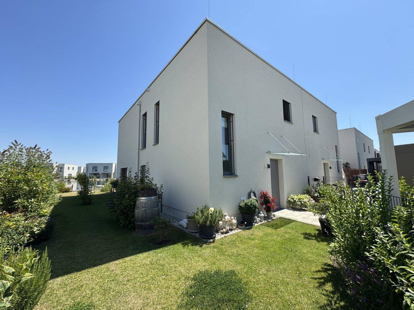 Moderne Hausfassade mit schrägem Dach, umgeben von einem grünen Garten und blühenden Pflanzen.