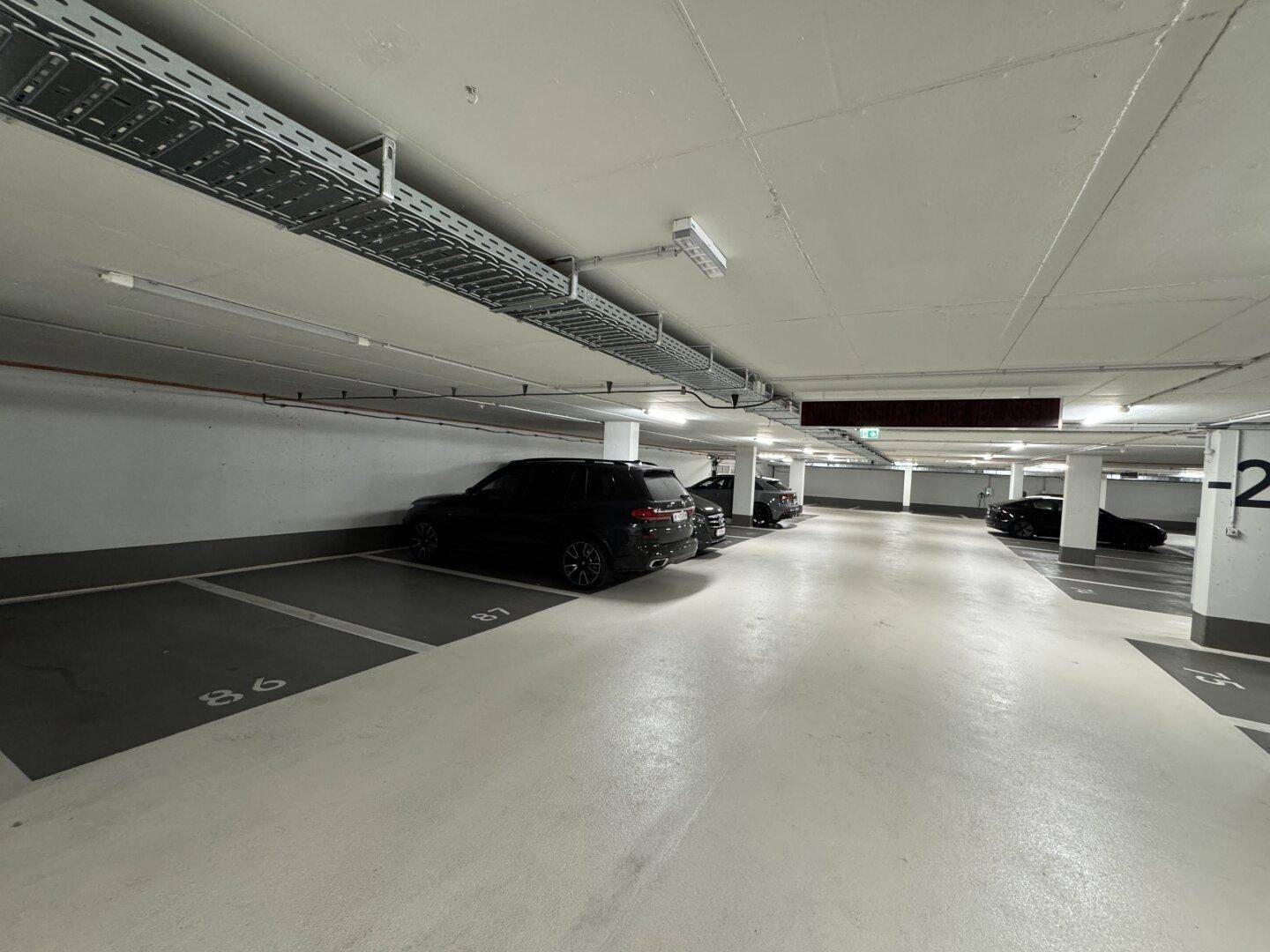 Weitläufige Tiefgarage mit mehreren Parkplätzen, sichtbaren Leitungen an der Decke und guter Beleuchtung.
