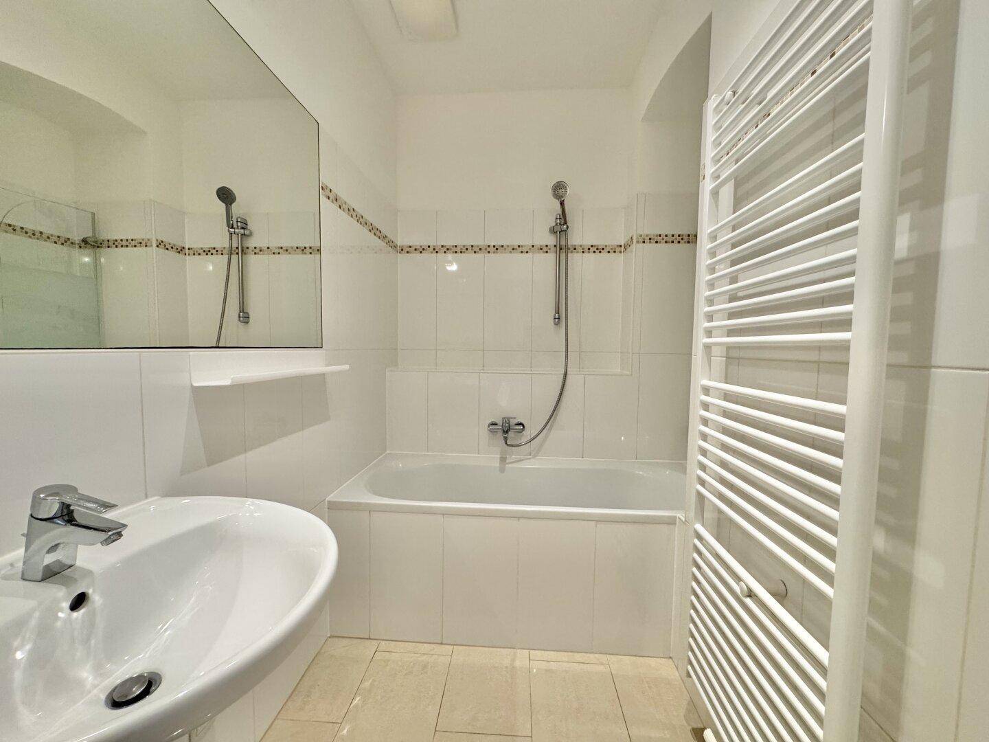 Helles Badezimmer mit Badewanne, Waschbecken und beheiztem Handtuchhalter, komplett gefliest.