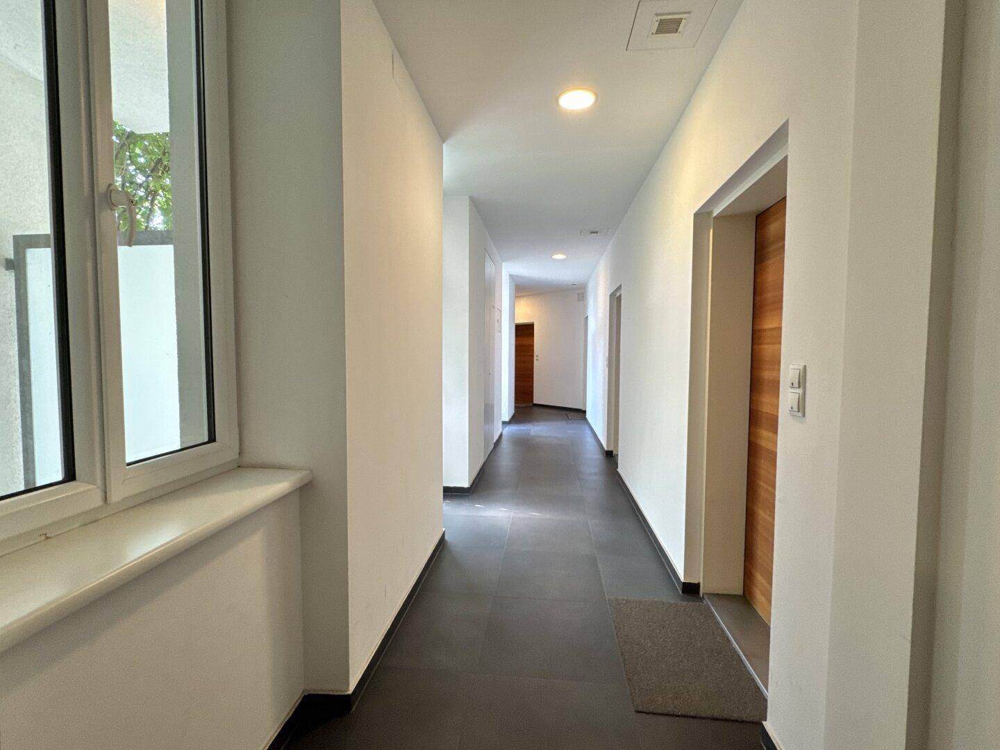 Langer, heller Flur mit dunklem Fliesenboden und Fenstern, führt zu verschiedenen Zimmern.