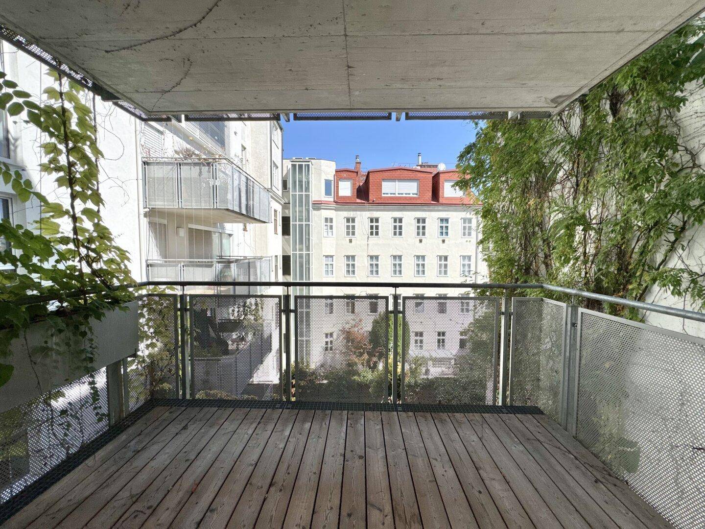 Balkon mit Holzboden und Metallgeländer bietet Blick auf die umliegenden Gebäude und Bäume.