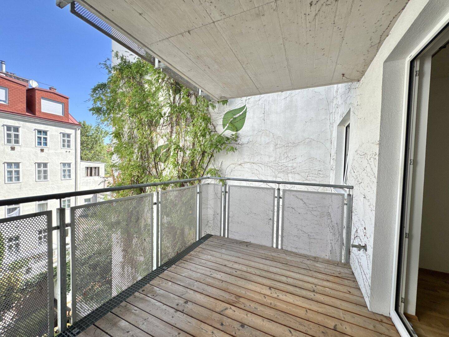 Sonniger Balkon mit Holzboden und Blick auf die begrünte Fassade und Nachbargebäude.
