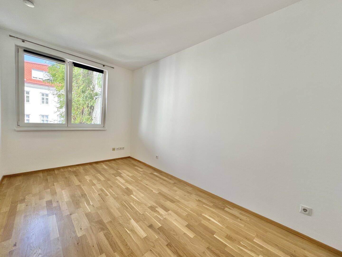 Freundliches Zimmer mit Parkettboden und großem Fenster, das viel Tageslicht hereinlässt.