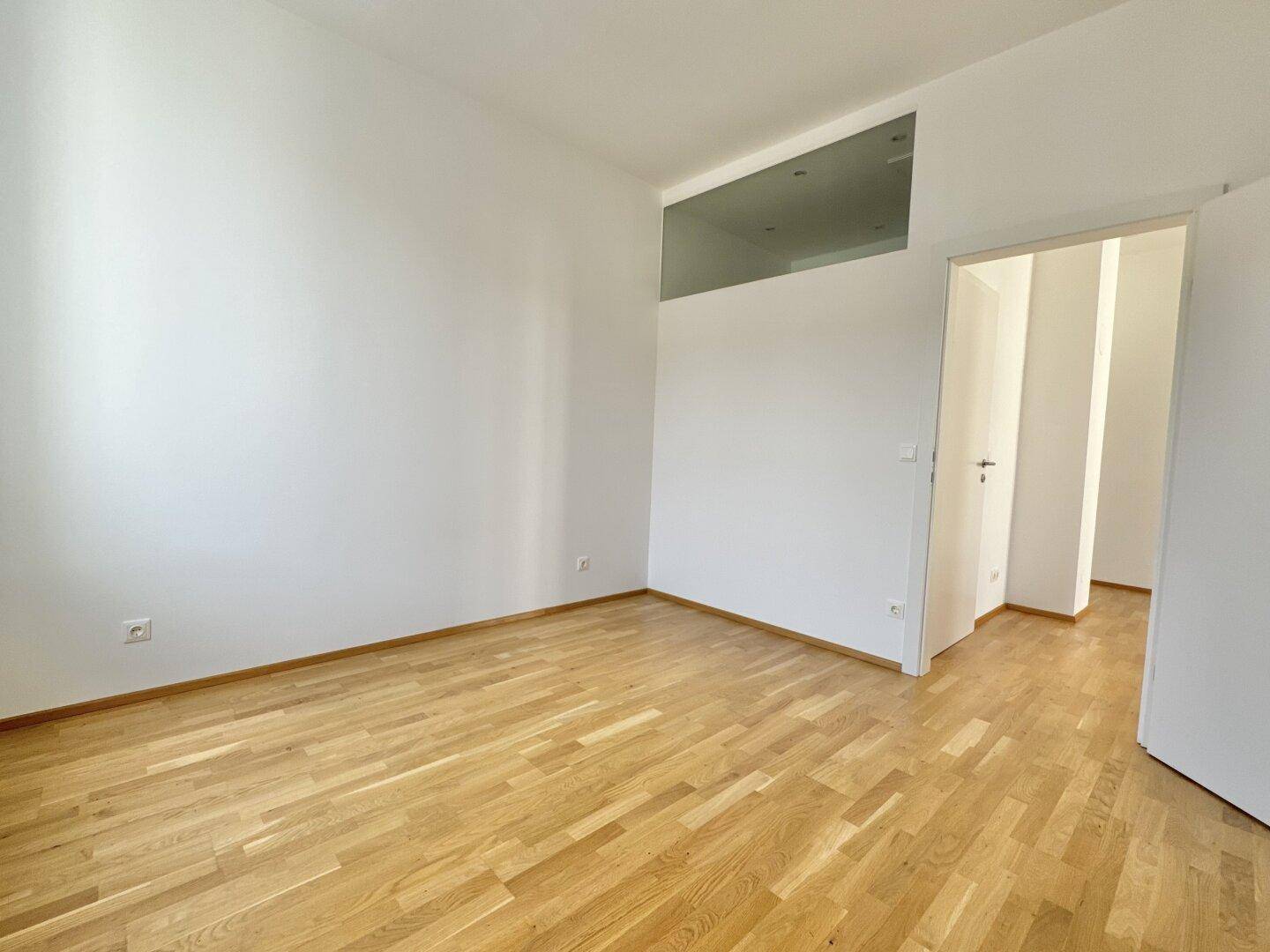 Leerer Raum mit Parkettboden und einer hohen Fensteröffnung über der Tür.