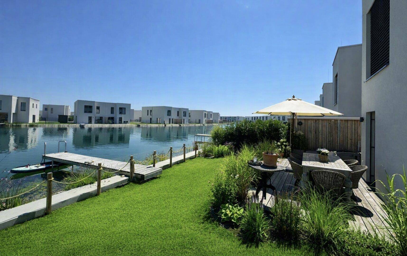Gemütliche Terrasse mit Sitzgelegenheiten und Sonnenschirm, direkt am See mit Blick auf die modernen Häuser.