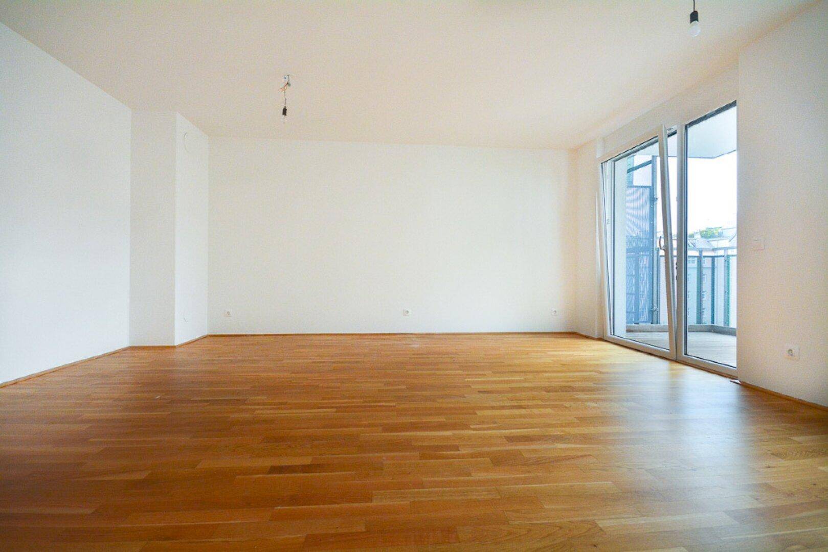 Leeres Wohnzimmer mit Holzboden und großen Fenstern, die viel Tageslicht hereinlassen.