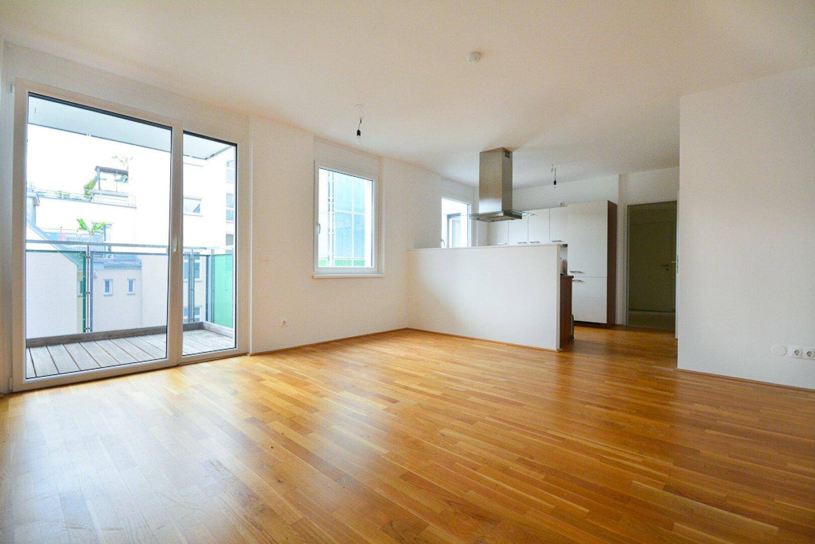 Geräumiges Wohnzimmer mit hellem Parkettboden und Zugang zu einem Balkon.