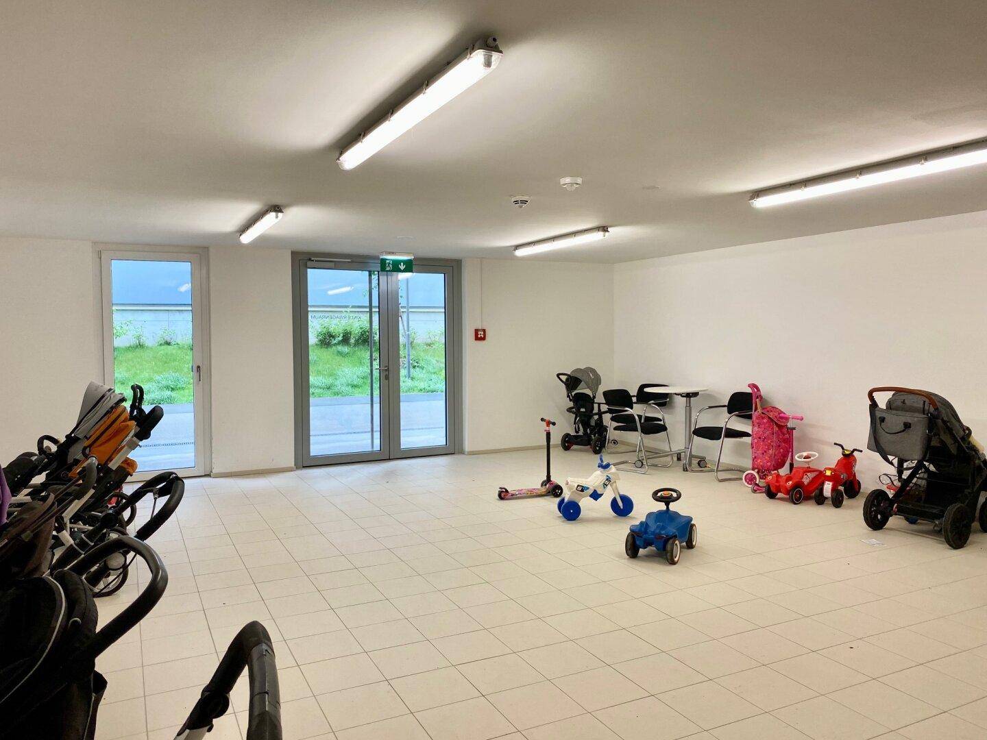 Heller Gemeinschaftsraum mit Fliesenboden, großen Fenstern und Abstellmöglichkeiten für Kinderwagen und Spielzeug.