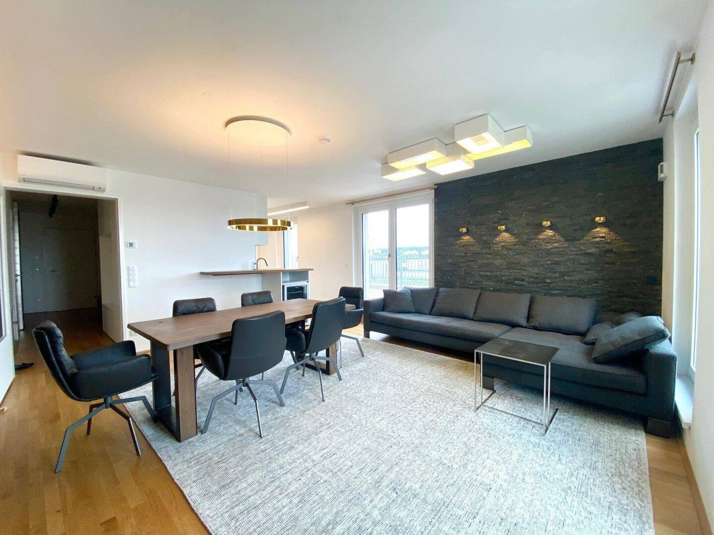 Moderner Wohn-Essbereich mit dunklem Sofa, Esstisch und offener Küchenzeile.