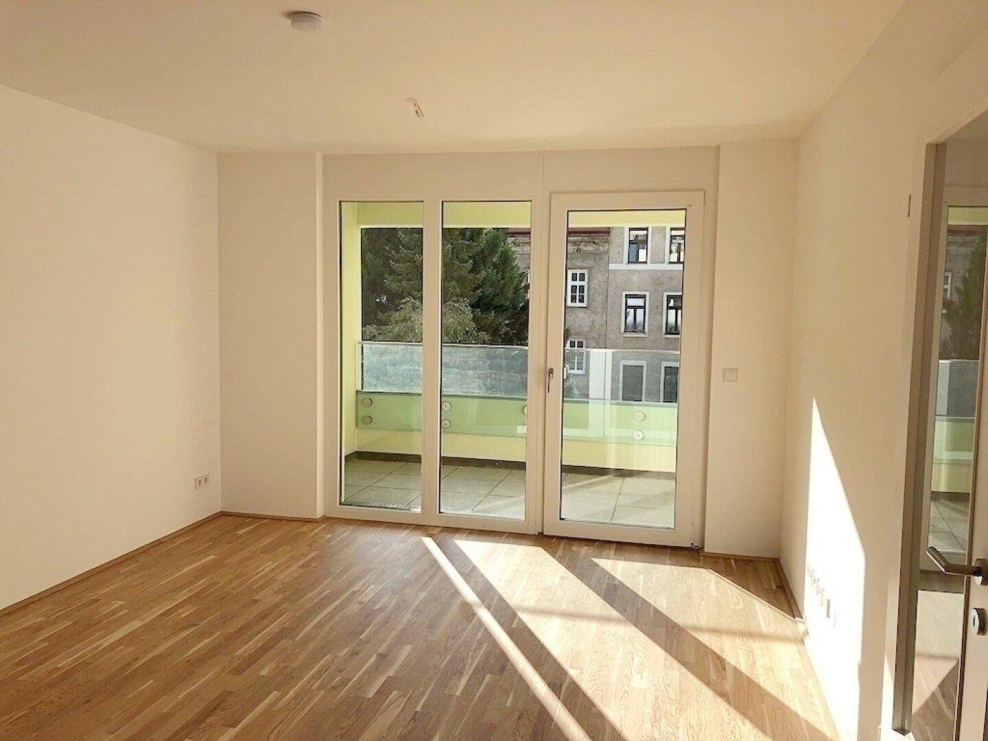 Helles Wohnzimmer mit Parkettboden und Zugang zu einem Balkon, durchflutet von Sonnenlicht.