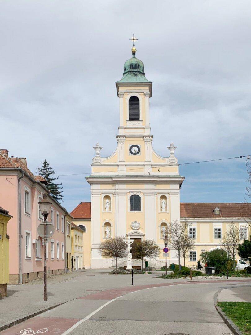 Historische Kirche mit barocker Fassade und einem Platz mit Bäumen in einer charmanten Stadtansicht.