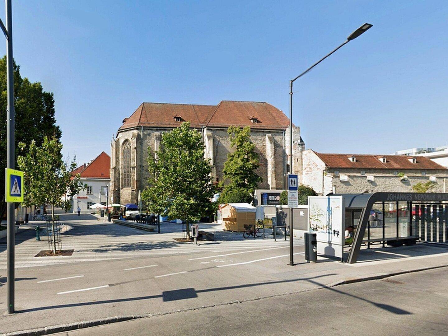 Historische Kirche und belebte Straße mit Bushaltestelle und Bäumen an einem sonnigen Tag.